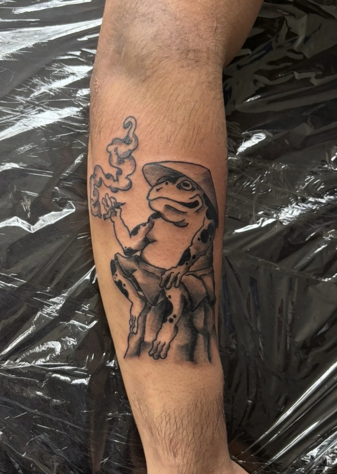 Tattoo of a frog with a Japanese style hat smoking with smoke rising from its finger, sitting on a rock.
