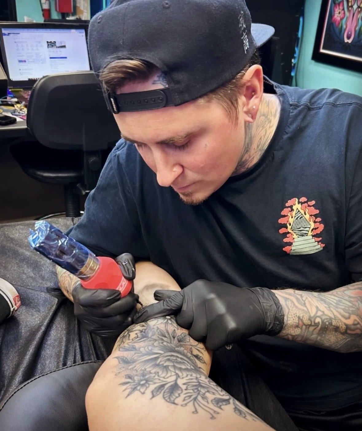 A man tattooing a person's arm in a tattoo parlor, with a tattoo machine in hand, wearing black gloves, a black cap worn backward, and a black T-shirt with a colorful design.