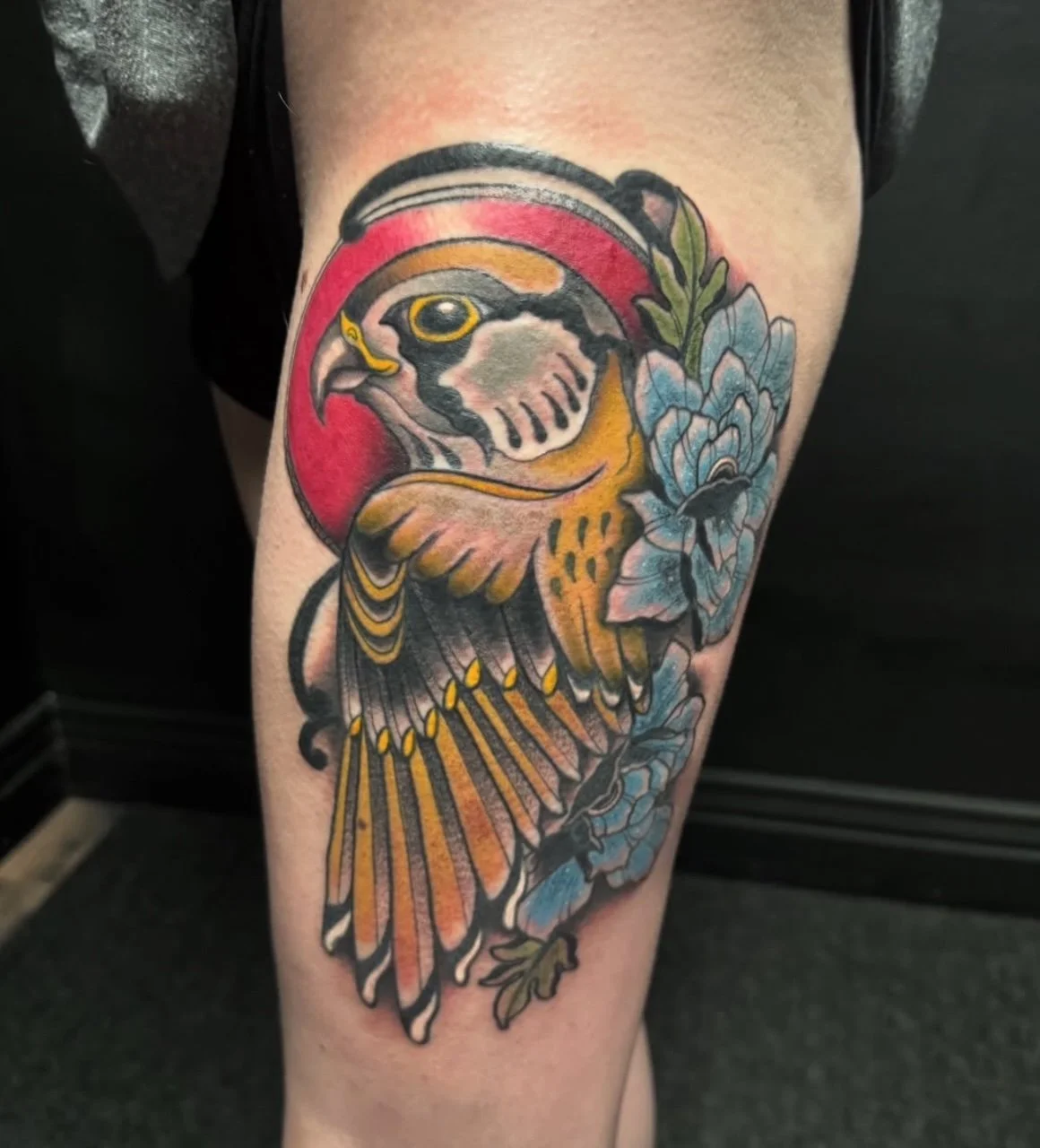 Colorful tattoo of a yellow bird with prominent wings, blue flowers, green leaves and touches of red on a person's thigh.