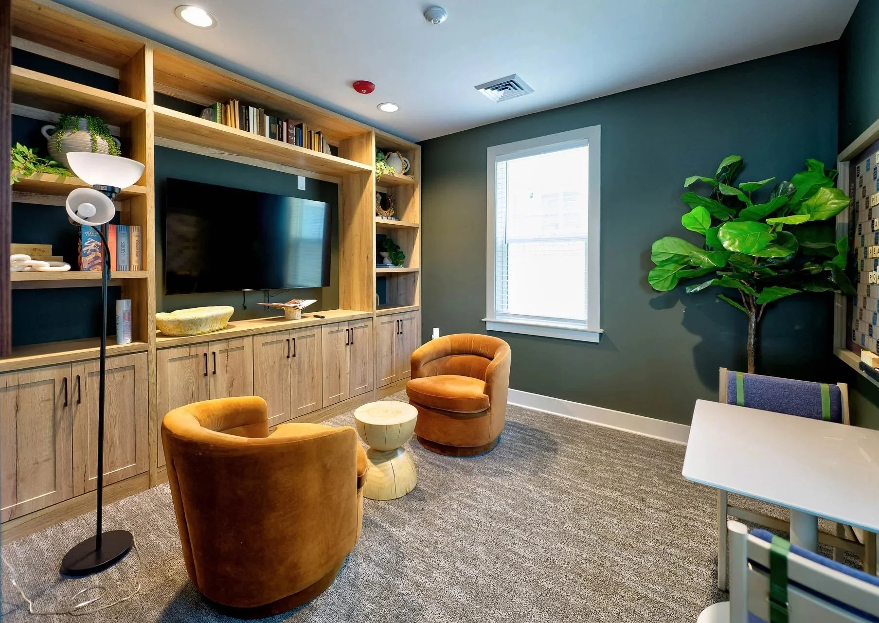 Interior of a room with a wall-mounted flat screen TV, wooden shelves with books and decorative items, a window with natural light, a tall green plant, orange velvet chairs, a white side table, and a small white table with multicolored chairs.