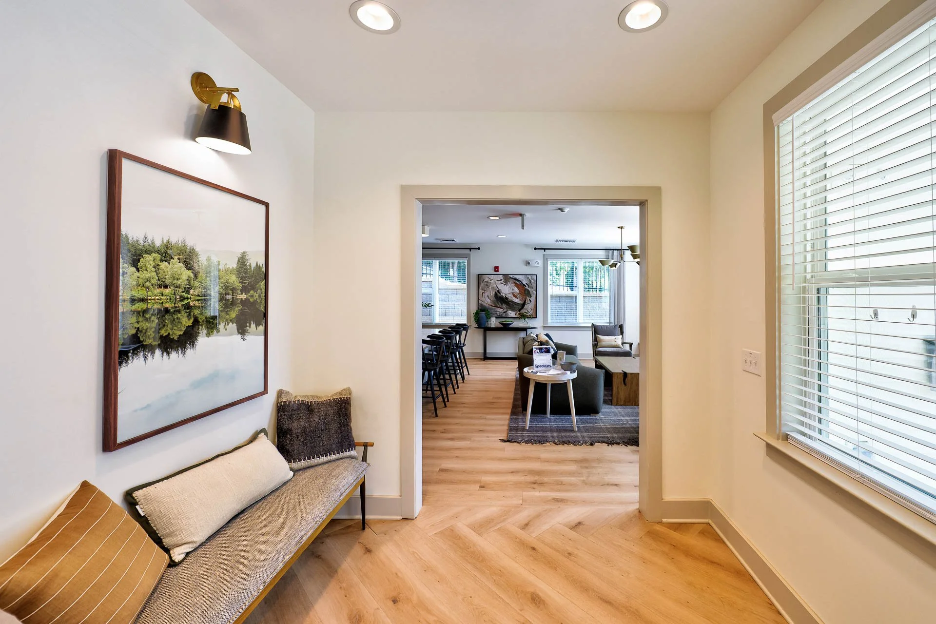 Interior view of a modern living room with a painted white wall, a framed landscape photograph, a brown bench with pillows, and large windows with blinds. Through a doorway, a second room features additional seating, artwork, and windows.