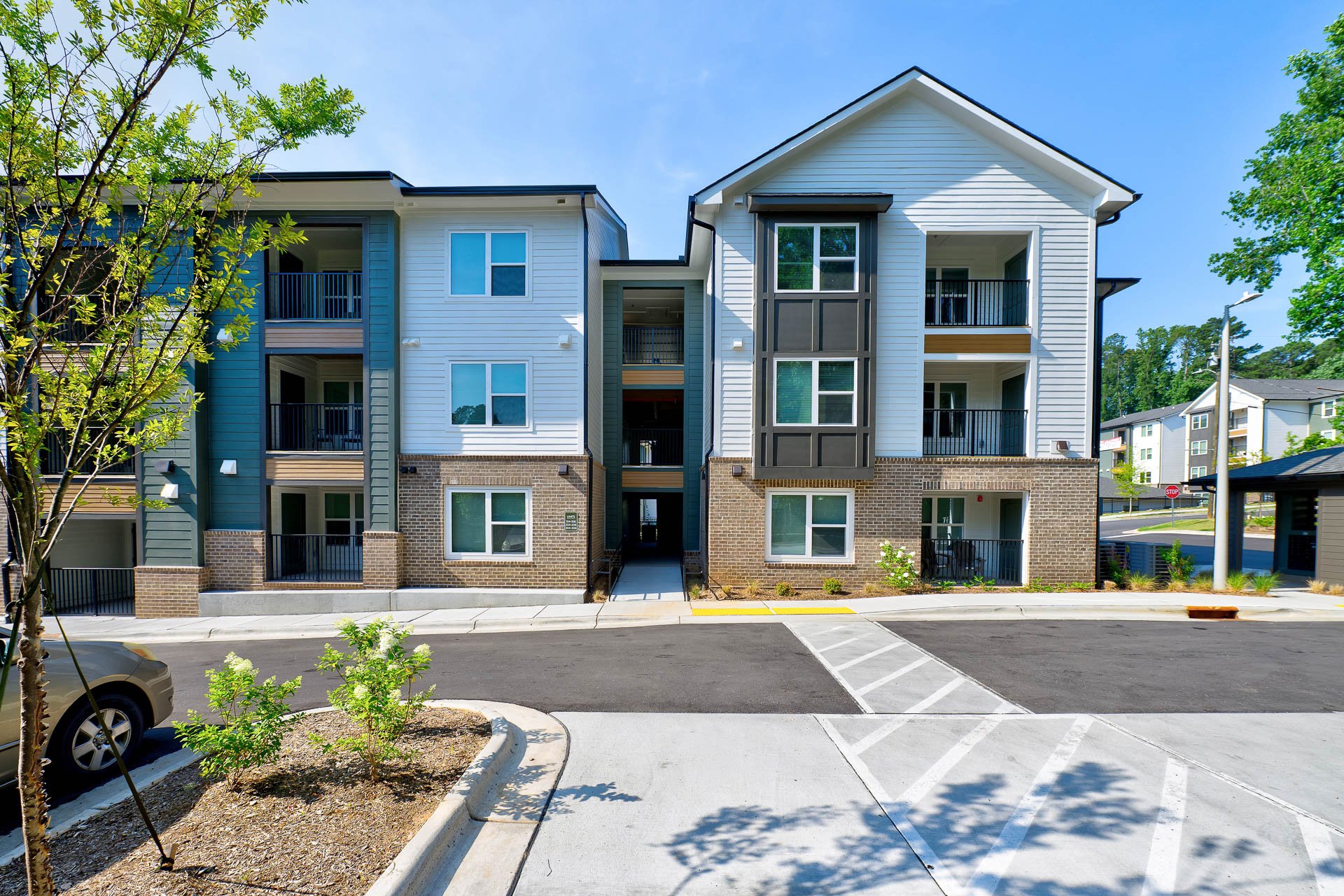 Modern multi-story residential building with white siding, brick accents, and balconies, under a clear blue sky at The Pines at Peach Road in Raleigh, NC.