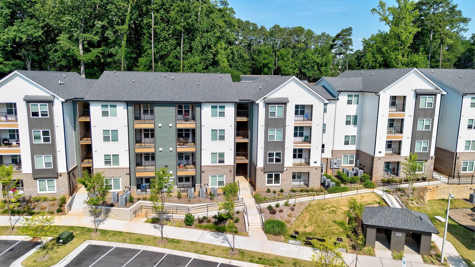 A modern multi-story apartment building with balconies, surrounded by trees and a landscaped yard, under a clear blue sky.