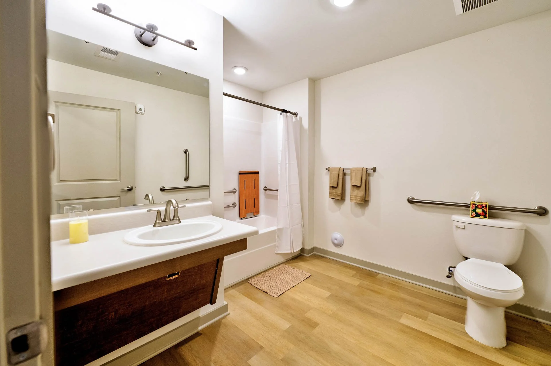 A clean, modern bathroom with a white sink, wooden vanity, and a large mirror. The bathroom has wood flooring, beige towels on a towel rack, a white shower curtain, and a toilet with a small decorative item on top. The ceiling has recessed lighting a