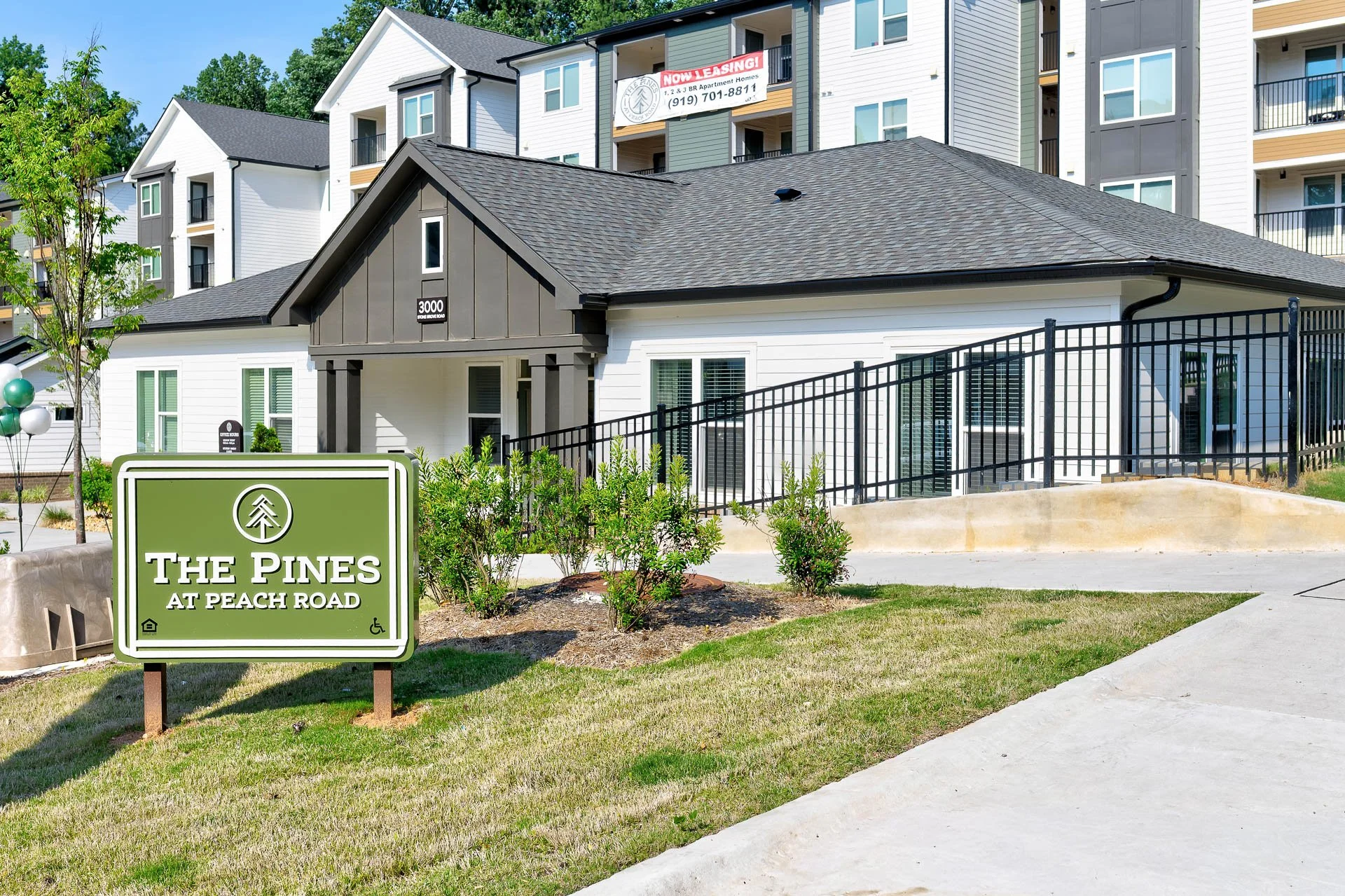 Apartment complex with a sign reading 'The Pines at Peach Road', modern white and gray buildings with multiple floors and balconies, a small landscaped area with plants, balloons, and a pathway.