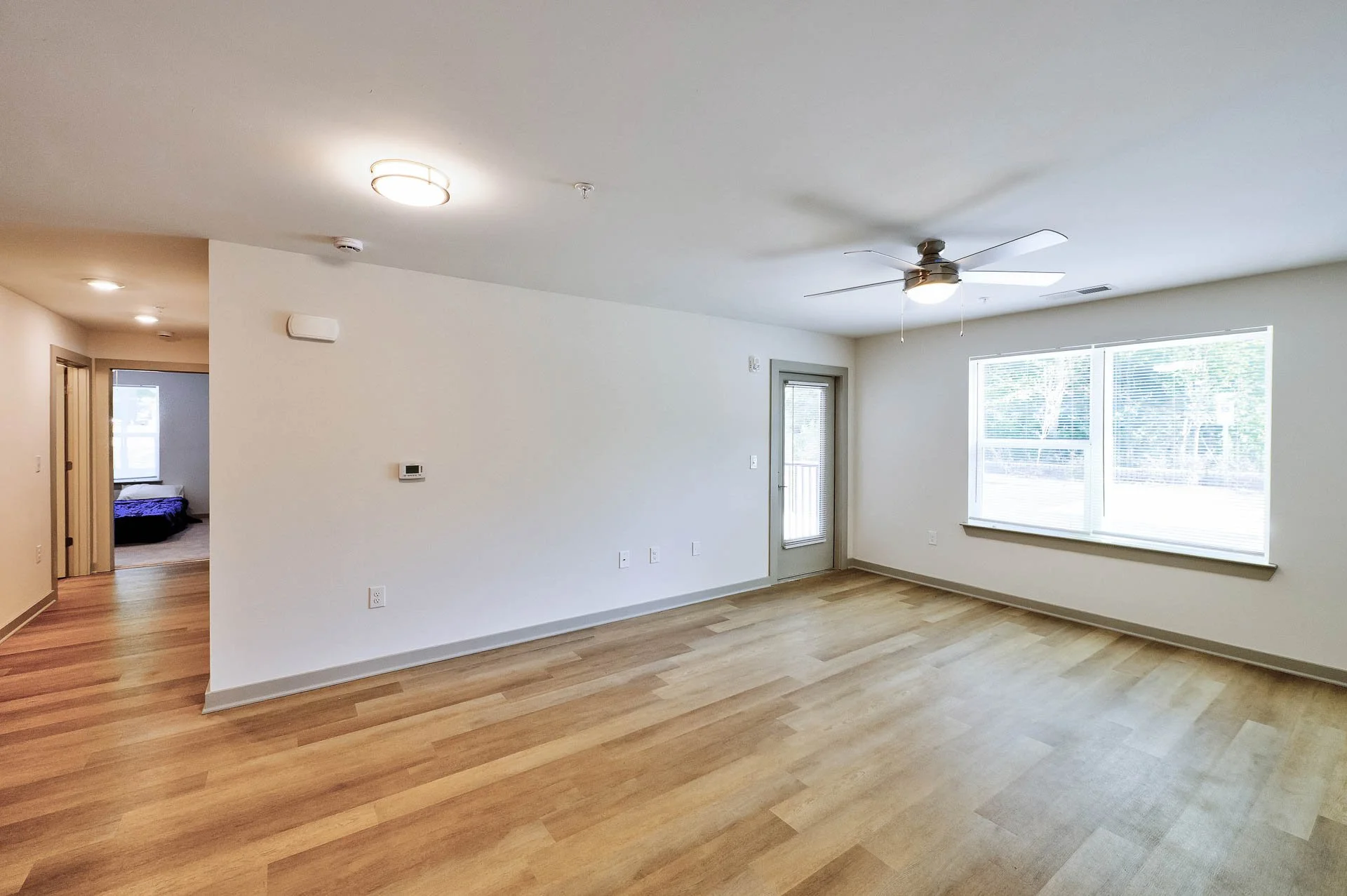 Empty living room with large window, ceiling fan, hardwood floors, and white walls, with a view into a bedroom with a bed and window.