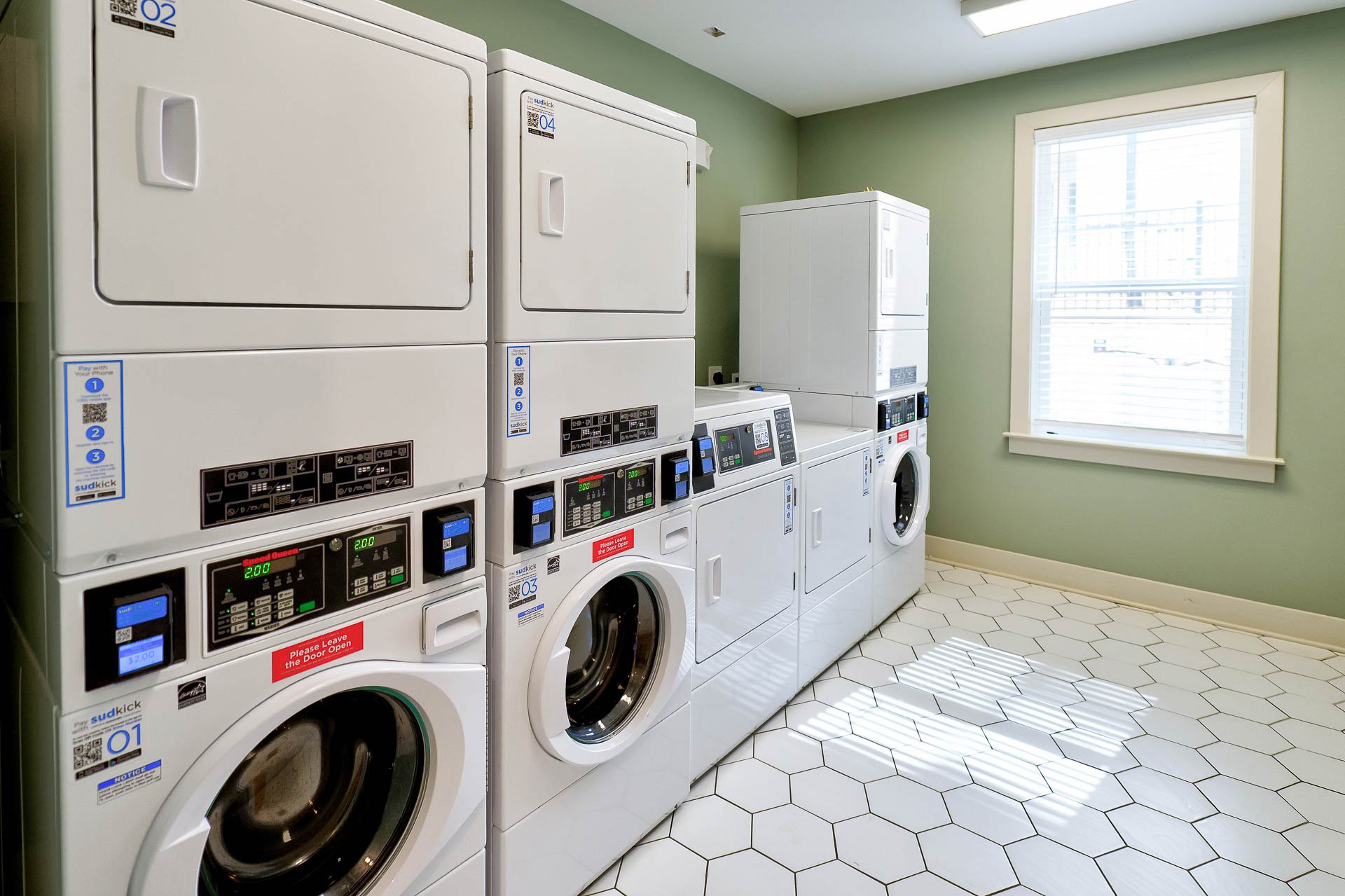 A laundry room with six white front-loading washing machines and dryers, arranged side by side. The room has light green walls, white hexagonal tiled floors, and a window on the right that lets in natural light.