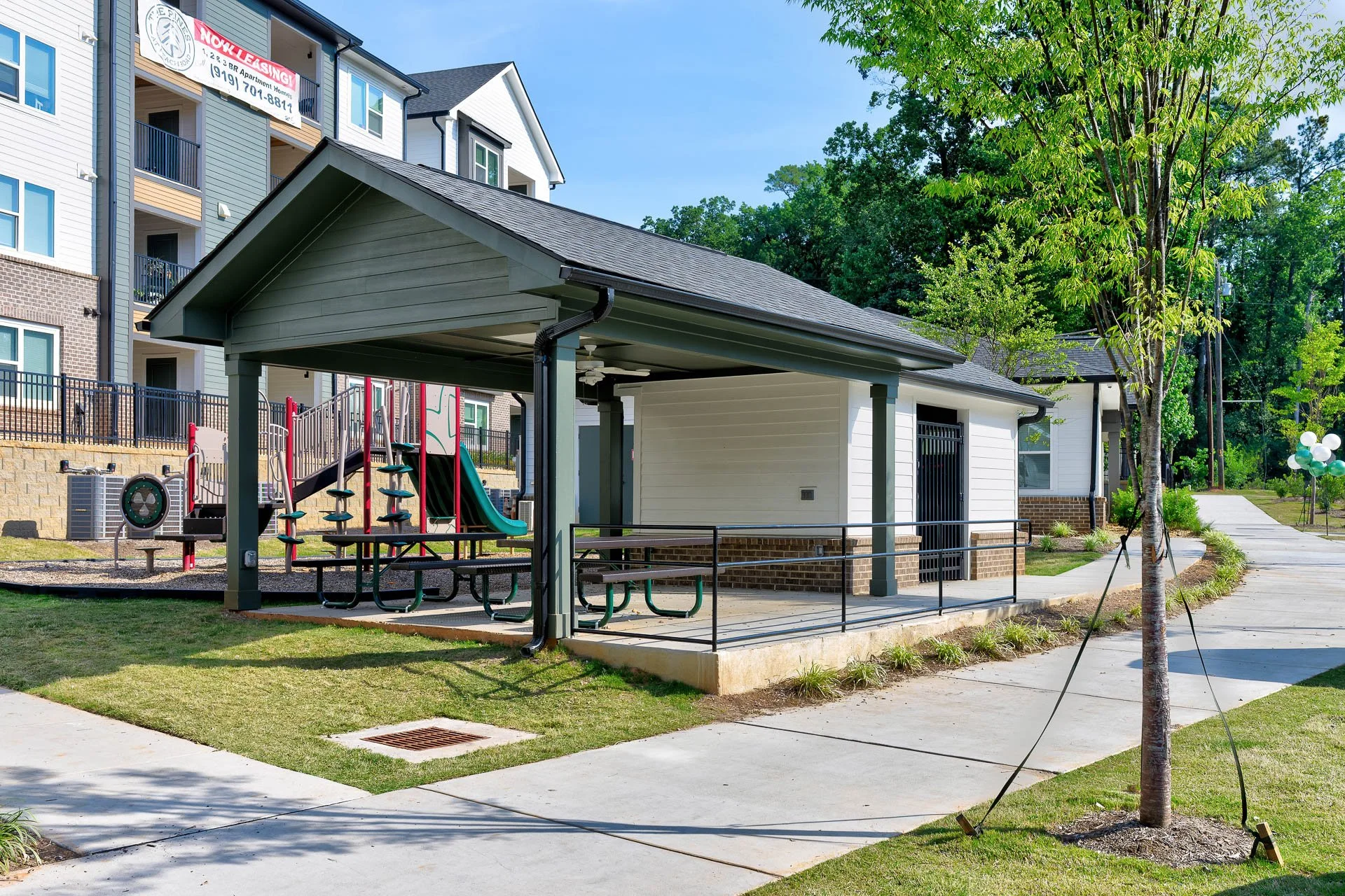 A community park area with a covered pavilion, playground equipment, sidewalk, trees, and surrounding residential apartment buildings under a clear blue sky.