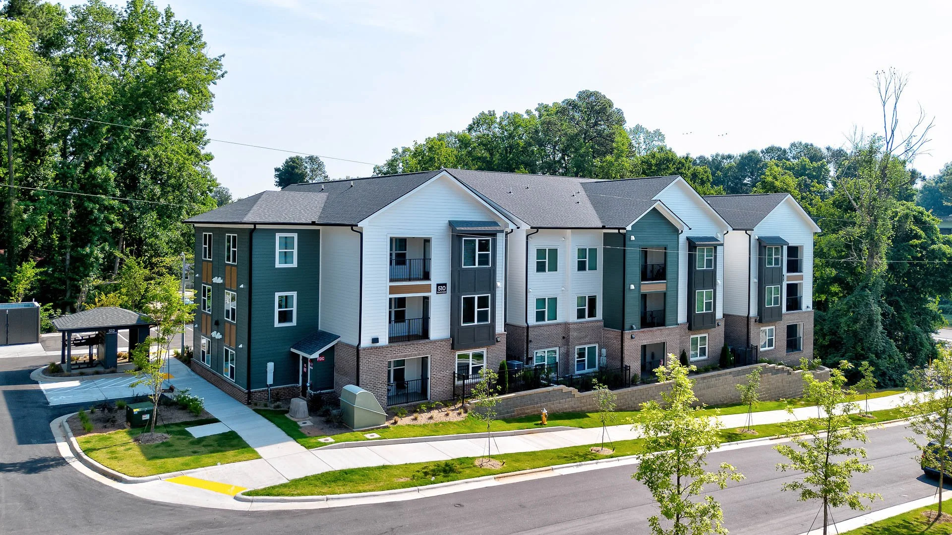 Newly constructed residential apartment building with multiple floors, parking lot, sidewalk, landscaped grass, young trees, and wooded area in background at The Pines at Peach Road in Raleigh, NC.