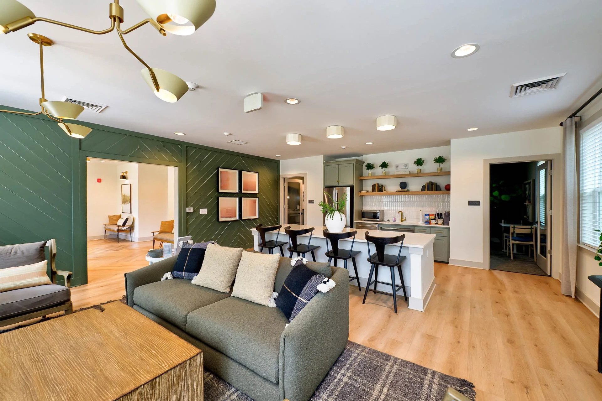 Open-concept living room with green accent wall, beige sofa with throw pillows, wooden coffee table, and kitchen with bar seating, plants, and modern decor.