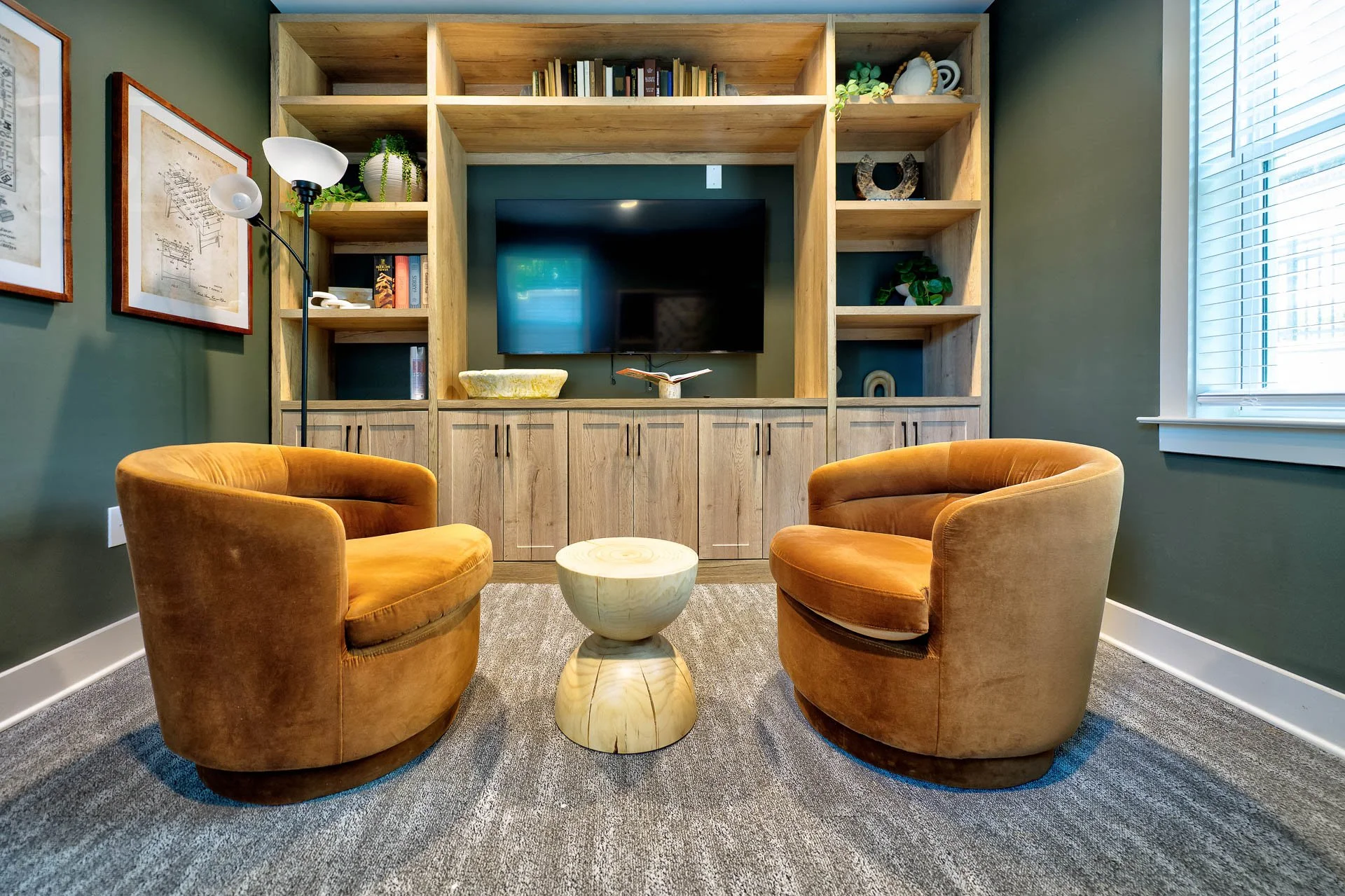 Living room with two orange velvet armchairs facing each other, a small light-colored wooden side table between them, a gray carpet, a large wall-mounted flat-screen TV, built-in wooden shelves with books, plants, and decorative objects, green walls,
