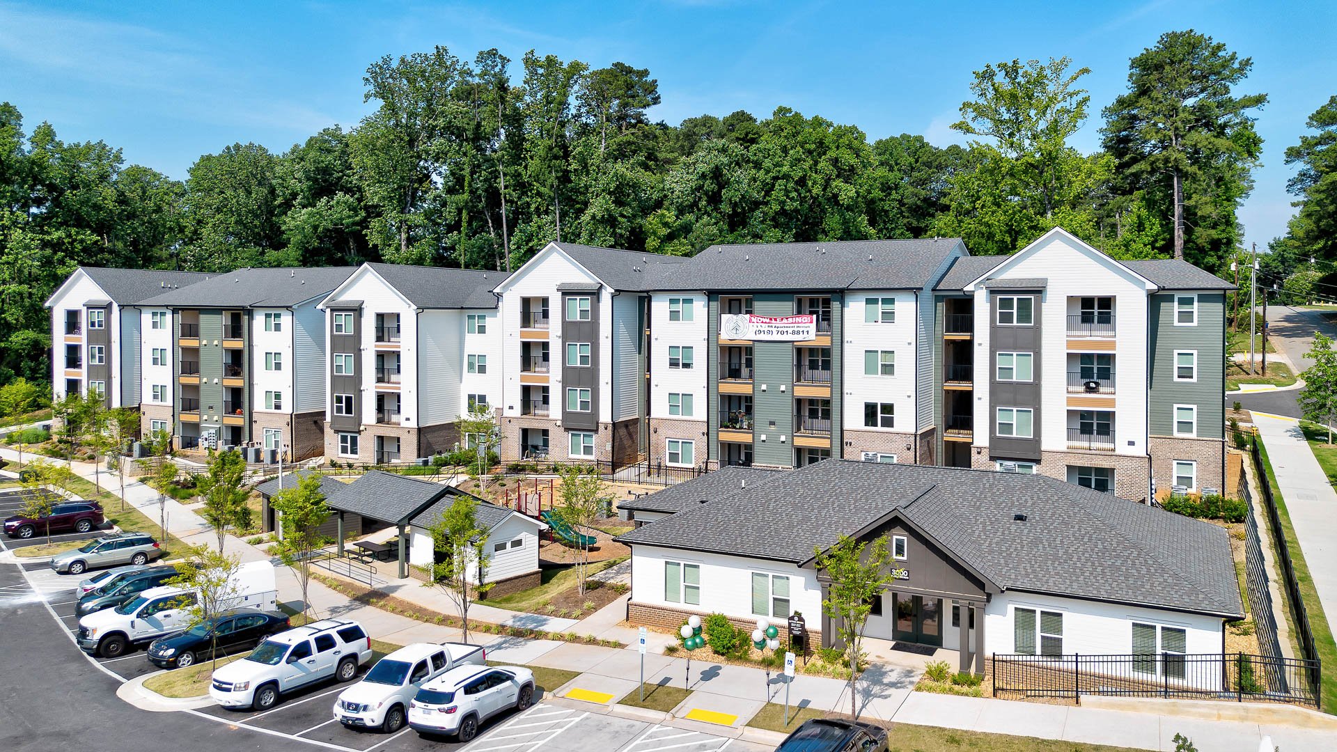 Multiple residential apartment buildings with a parking lot and green trees in the background at The Pines at Peach Road in Raleigh, NC.