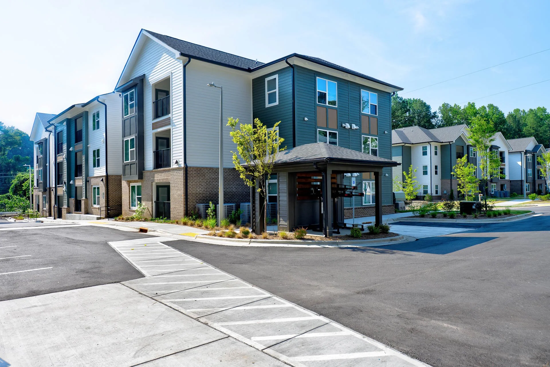 View of a modern apartment complex with multiple stories, painted in white and blue, with small trees planted along the sidewalk, and a parking lot with empty spaces in the foreground at The Pines at Peach Road in Raleigh, NC.