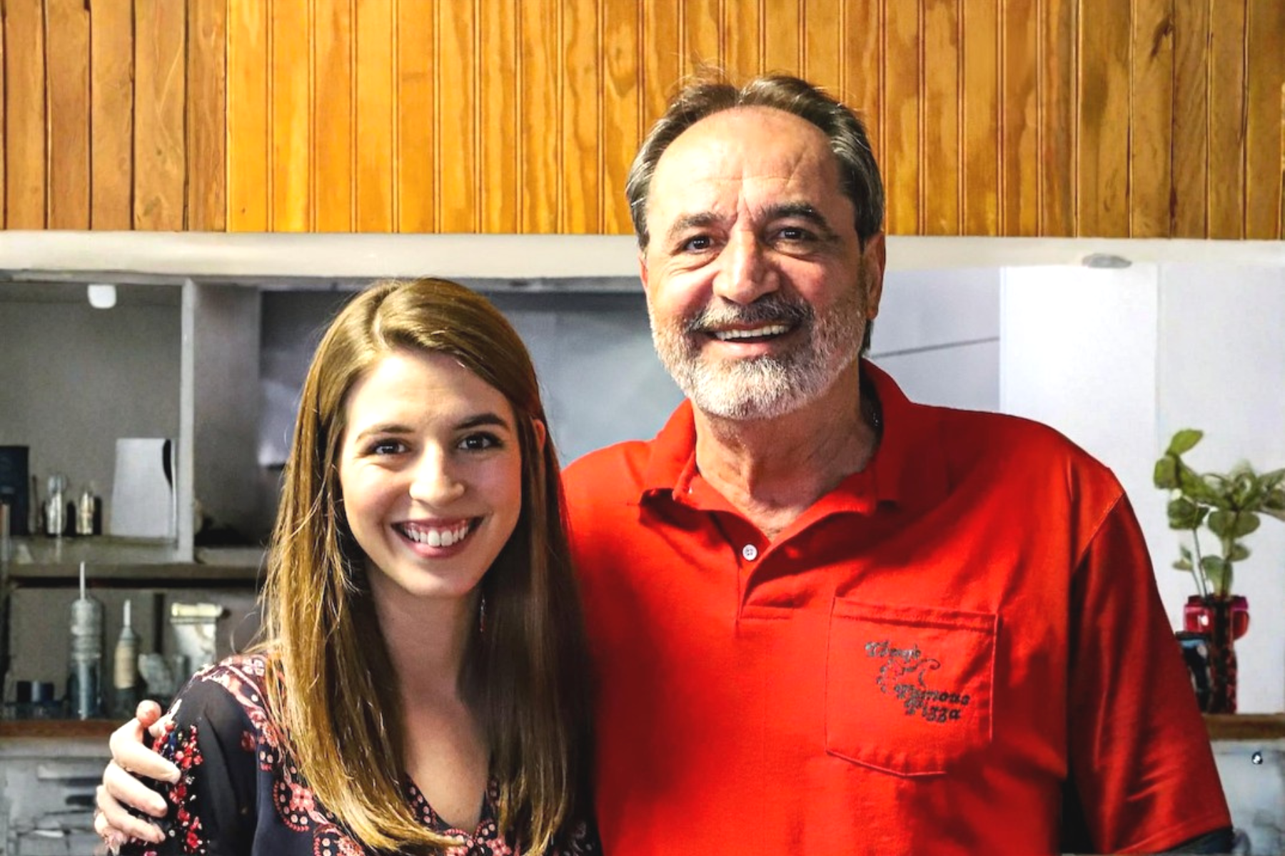 A young woman and an older man smiling and posing together in a kitchen with wooden panel walls. The woman has long brown hair and is wearing a black dress with floral patterns. The man has gray hair, a beard, and is wearing a red shirt.