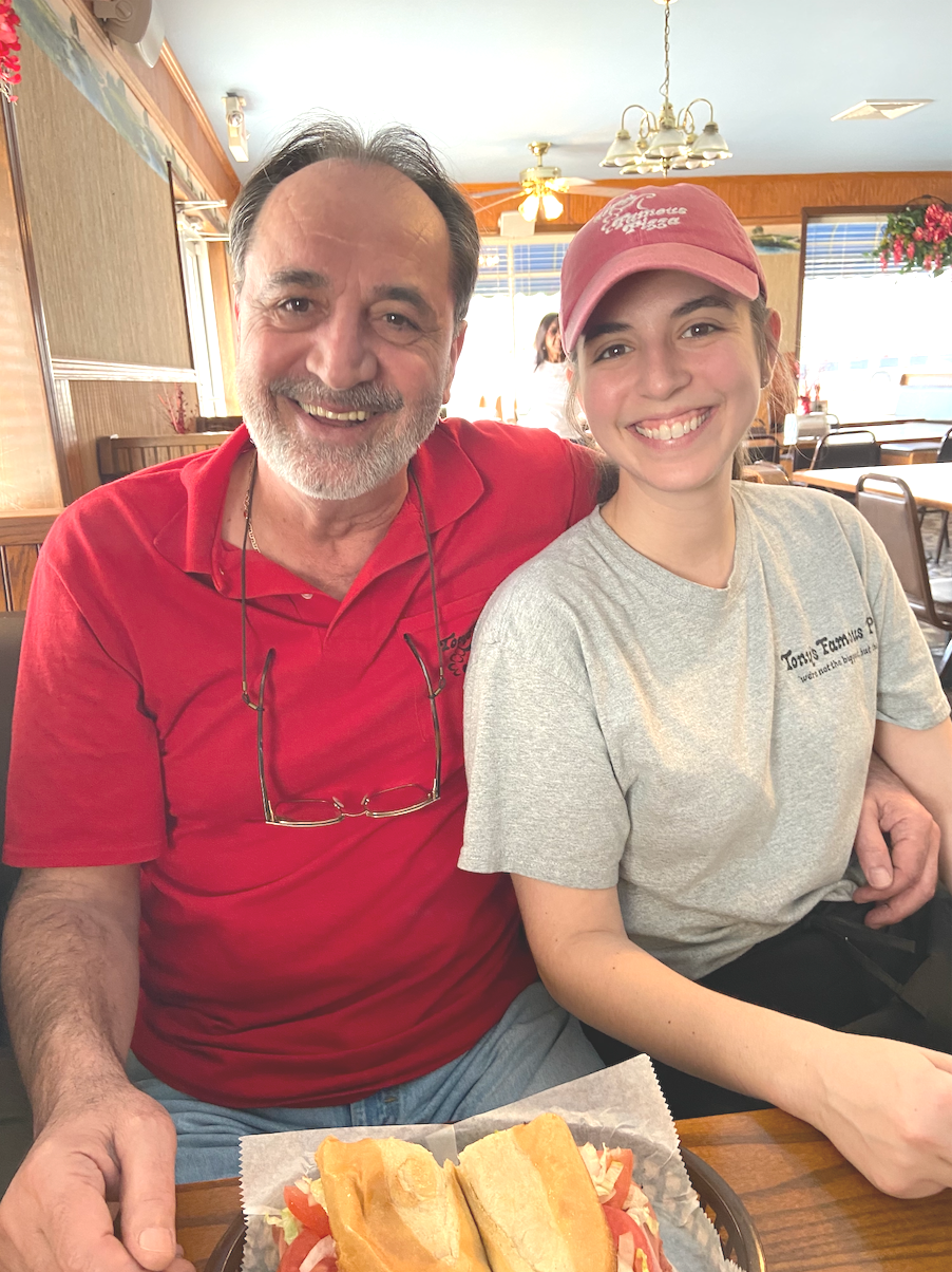 A smiling older man with a beard and glasses hanging from his collar, sitting next to a young woman in a pink cap and gray t-shirt, in a restaurant. There is a sandwich on the table in front of them.