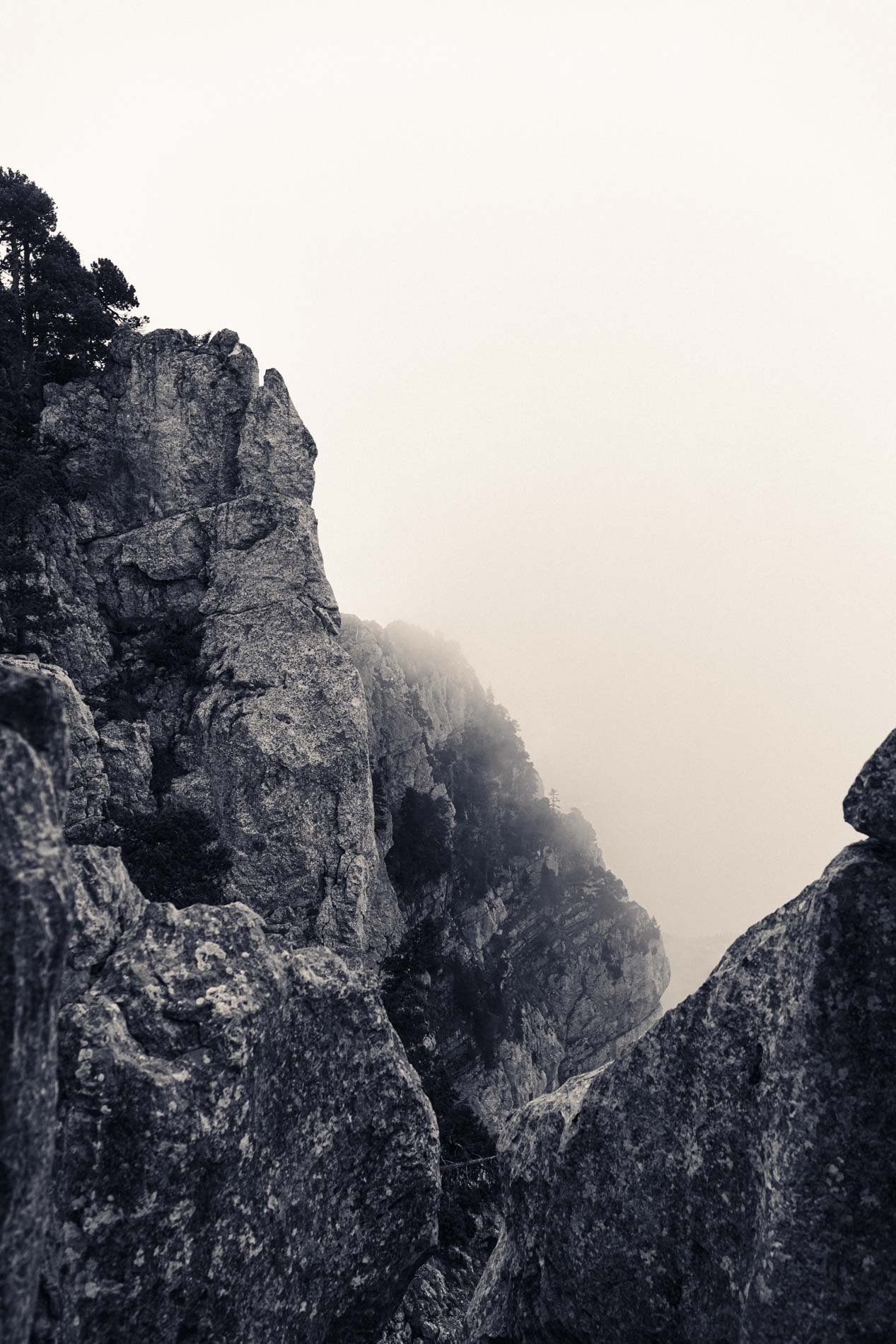 Paysage de montagnes rocheuses enveloppées de brume ou de nuages, vue de haut avec des rochers en premier plan.