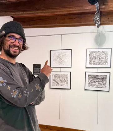 Un homme souriant avec lunettes, portant un bonnet, pointant vers des œuvres d'art abstraites encadrées sur un mur dans une galerie d'art.