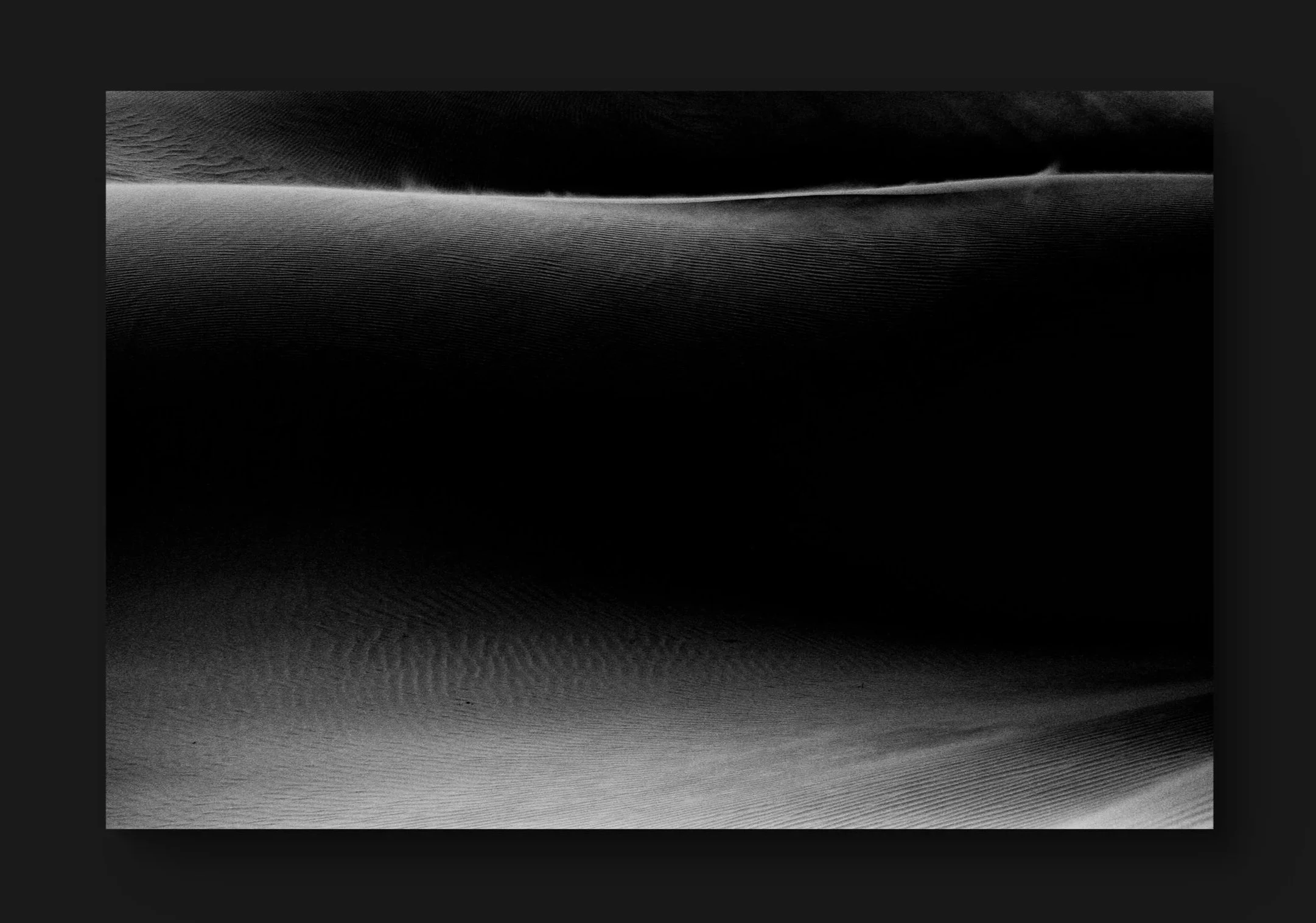 Une image noire et blanche représentant une dune de sable avec des motifs ondulés et une faible lumière créant des ombres contrastées.