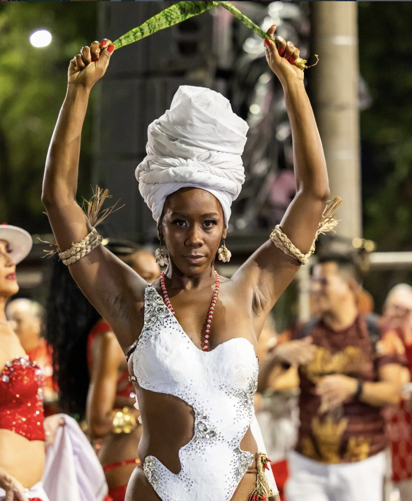 Sabrina Ginga com turbante branco e vestido branco decorado com pedrarias, participando de desfile do carnaval, rodeada por outras pessoas também vestidas com roupas tradicionais e coloridas.