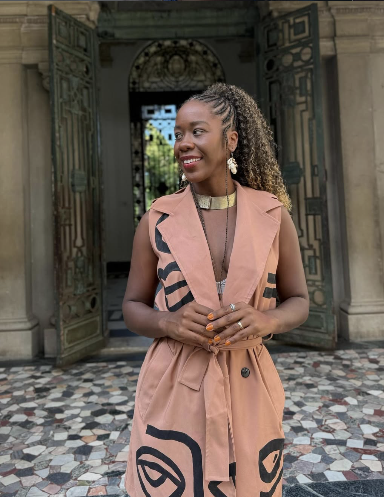 Sabrina Ginga sorridente com cabelo encaracolado segurando as mãos na frente, usando vestido sem mangas com detalhes negros, acessórios elegantes e brincos grandes, em frente a um portão de ferro antigo e detalhes arquitetônicos clássicos.