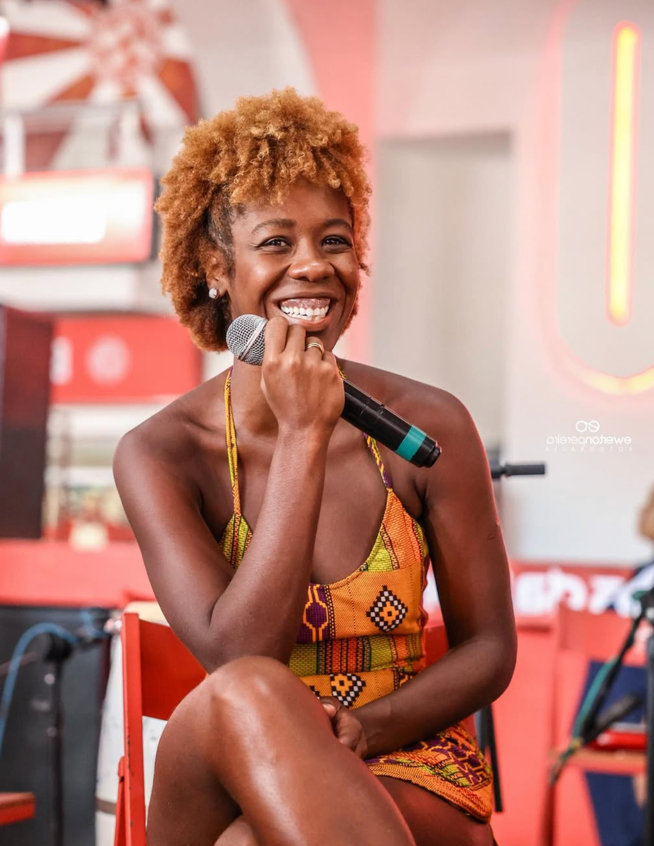 Sabrina Ginga sorridente segurando um microfone, vestida com roupa de tecido colorido com padrão africano, sentado em uma cadeira vermelha em um ambiente decorado com cores quentes e neon.