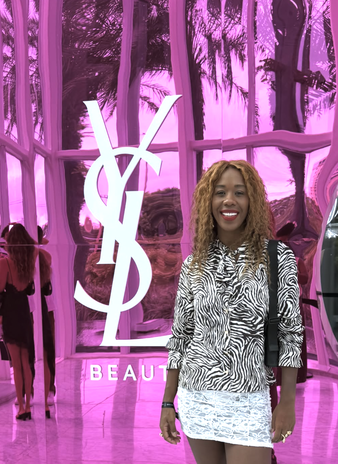 Sabrina Ginga sorridente com cabelo cacheado, vestindo blusa de zebra e saia branca, posando na frente de fundo rosa com logo de Yves Saint Laurent e árvores refletidas.
