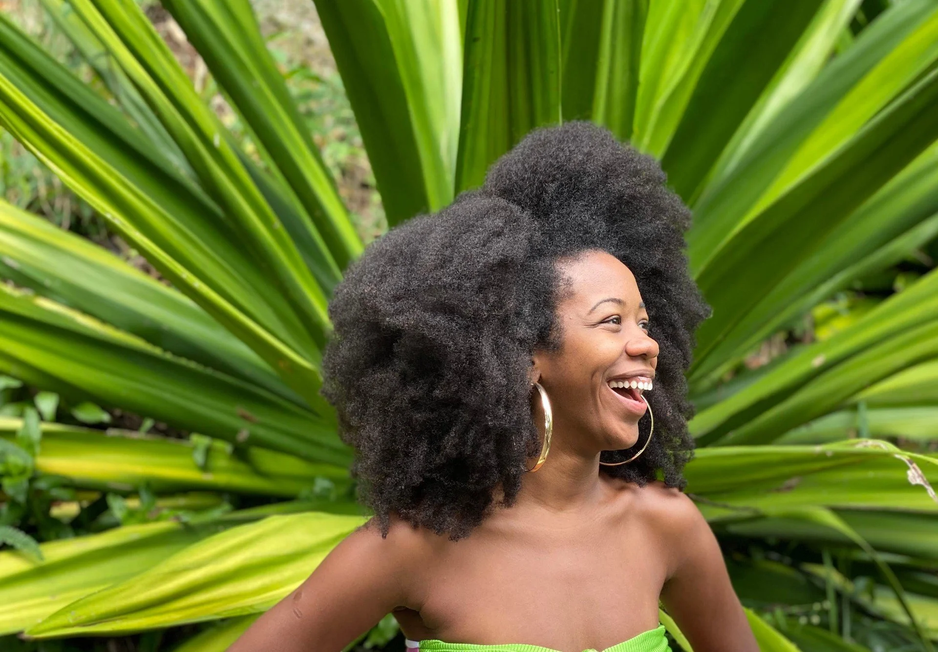 Sabrina Ginga sorridente com cabelo afro e brincos dourados, usando top verde, ao fundo folhas verdes.