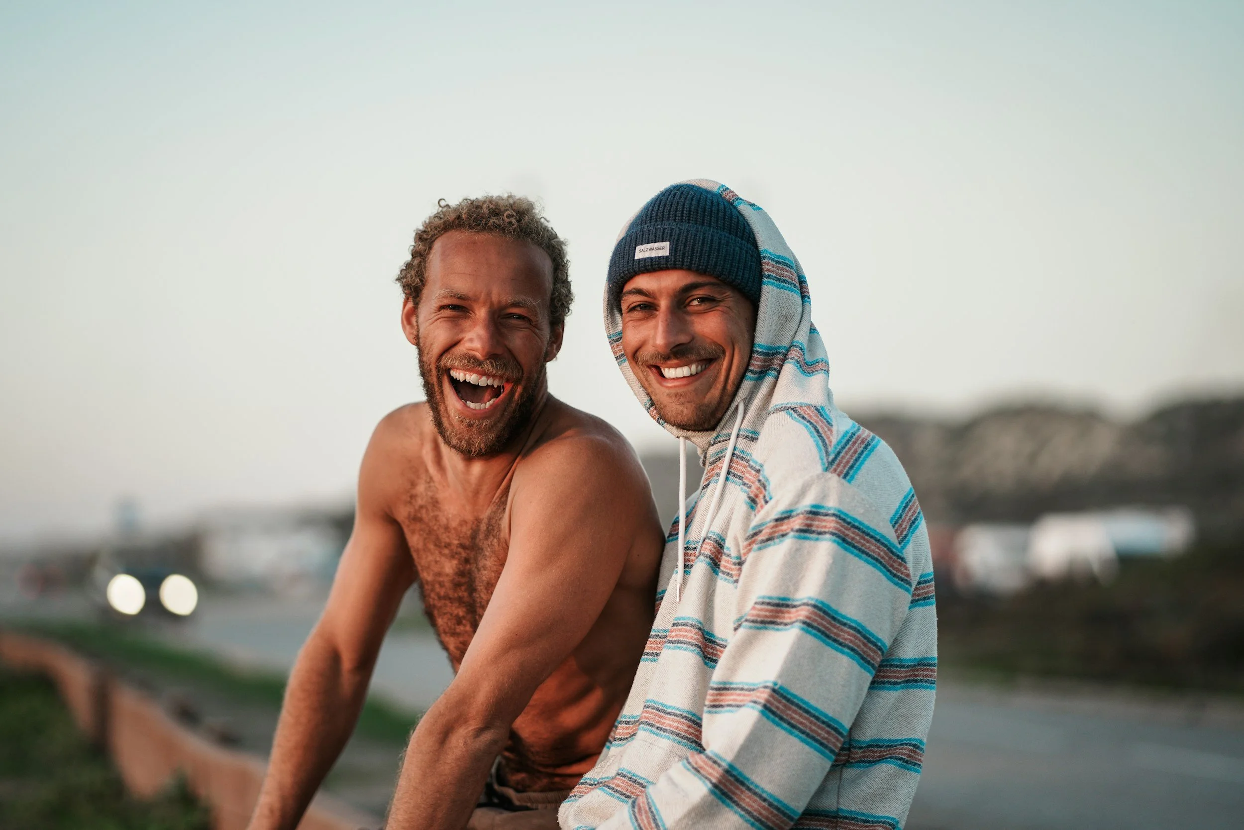 Two men smiling and sitting together outdoors, one wearing a striped hoodie and beanie, the other shirtless with curly hair.