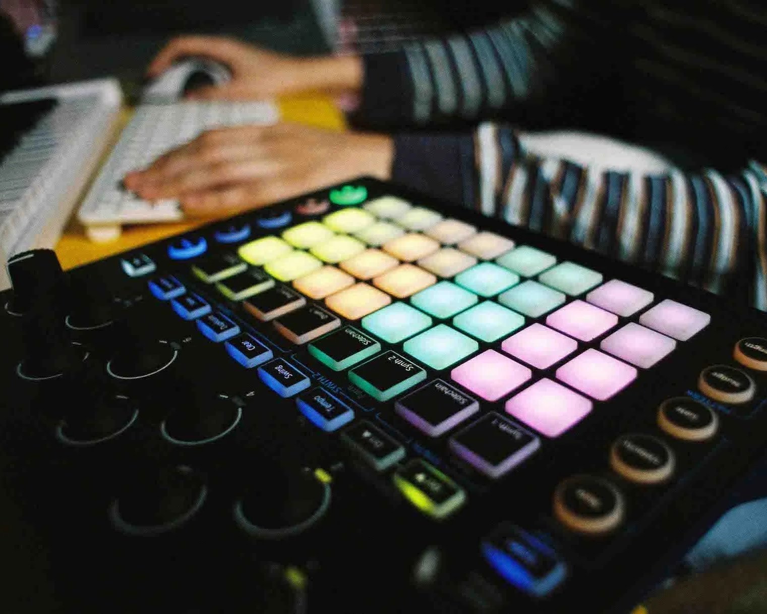 A person using a music production controller with colorful pads and knobs, and a laptop keyboard nearby.