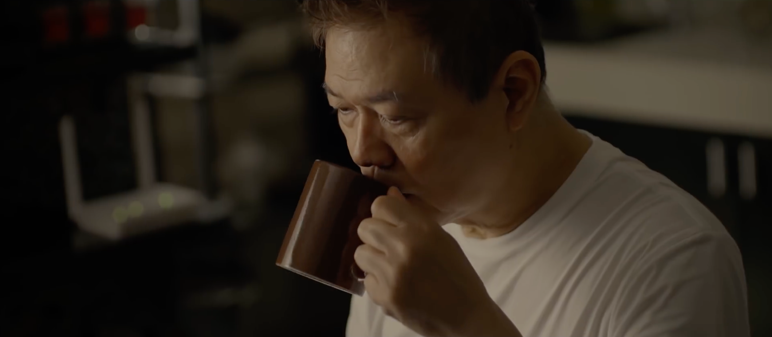 A man wearing a white shirt is sipping from a brown mug in a dimly lit room.