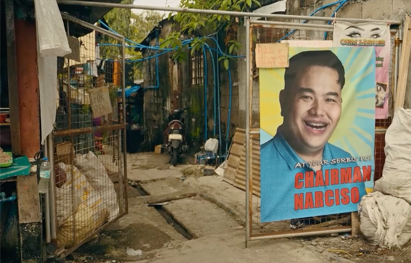 An outdoor scene with a metal fence displaying a campaign poster and signs. The poster features a smiling man with short black hair, wearing a blue shirt, with bold red text 'Chairman Narciso.' A small sign on the fence reads 'For Rent' and another a