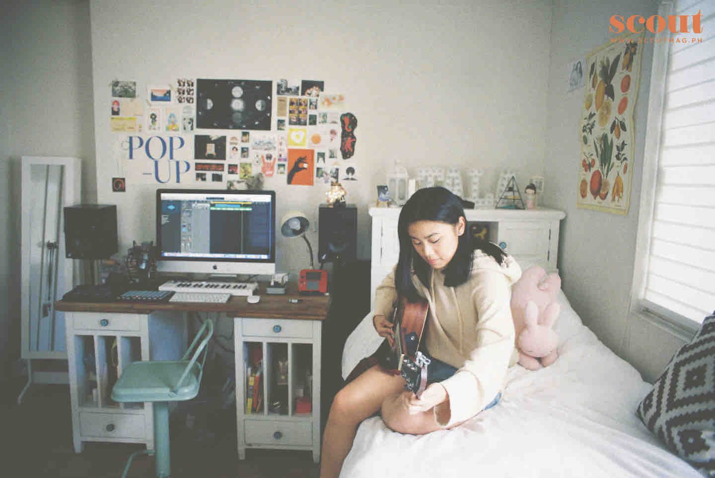 A girl sitting on a bed playing an acoustic guitar in a bedroom. The room has a desk with a computer, music equipment, and a wall decorated with various posters and pictures.