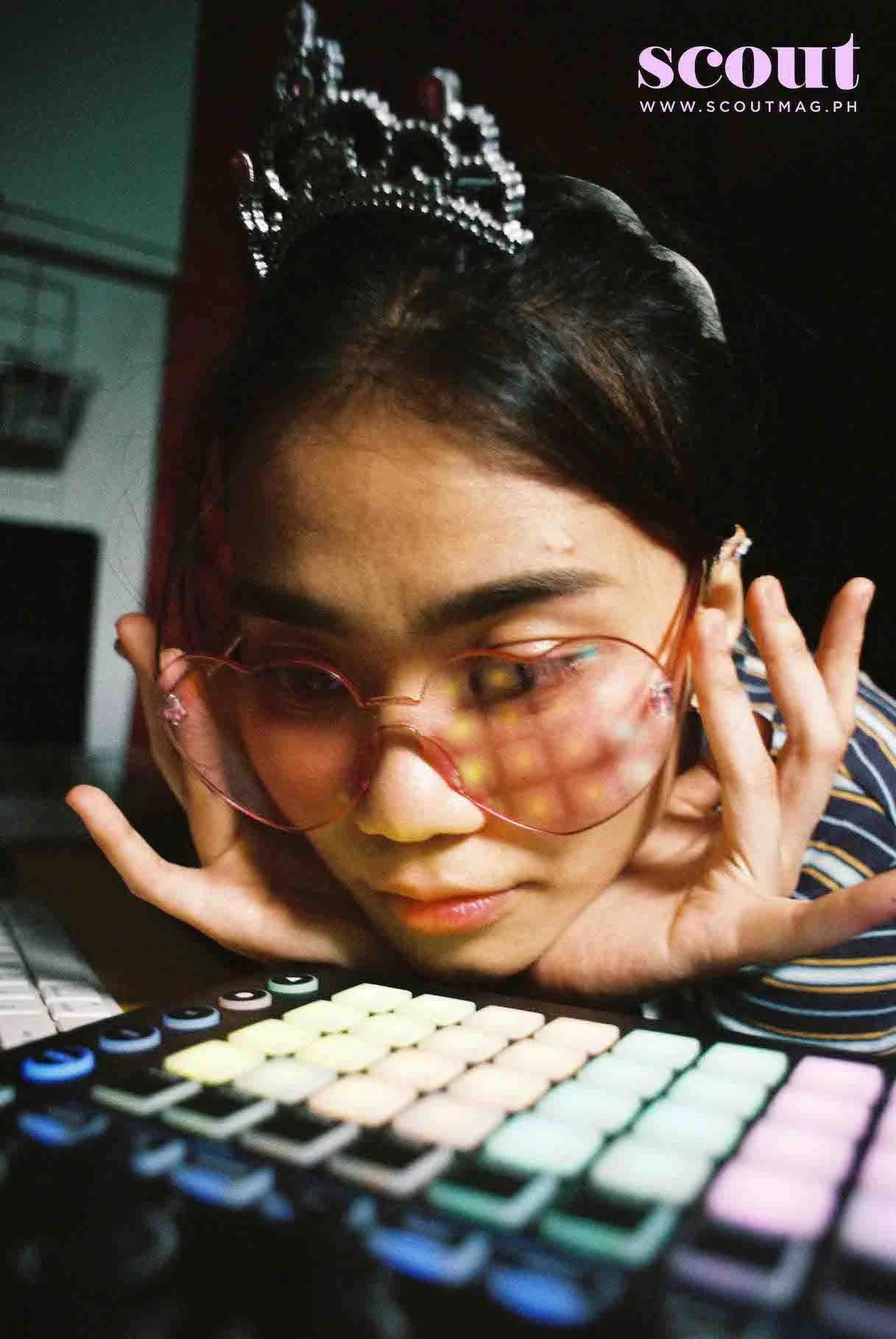 A young woman with dark hair and glasses rests her chin on her hand in front of a colorful keyboard. She wears a tiara and striped shirt, with a slightly blurred background.