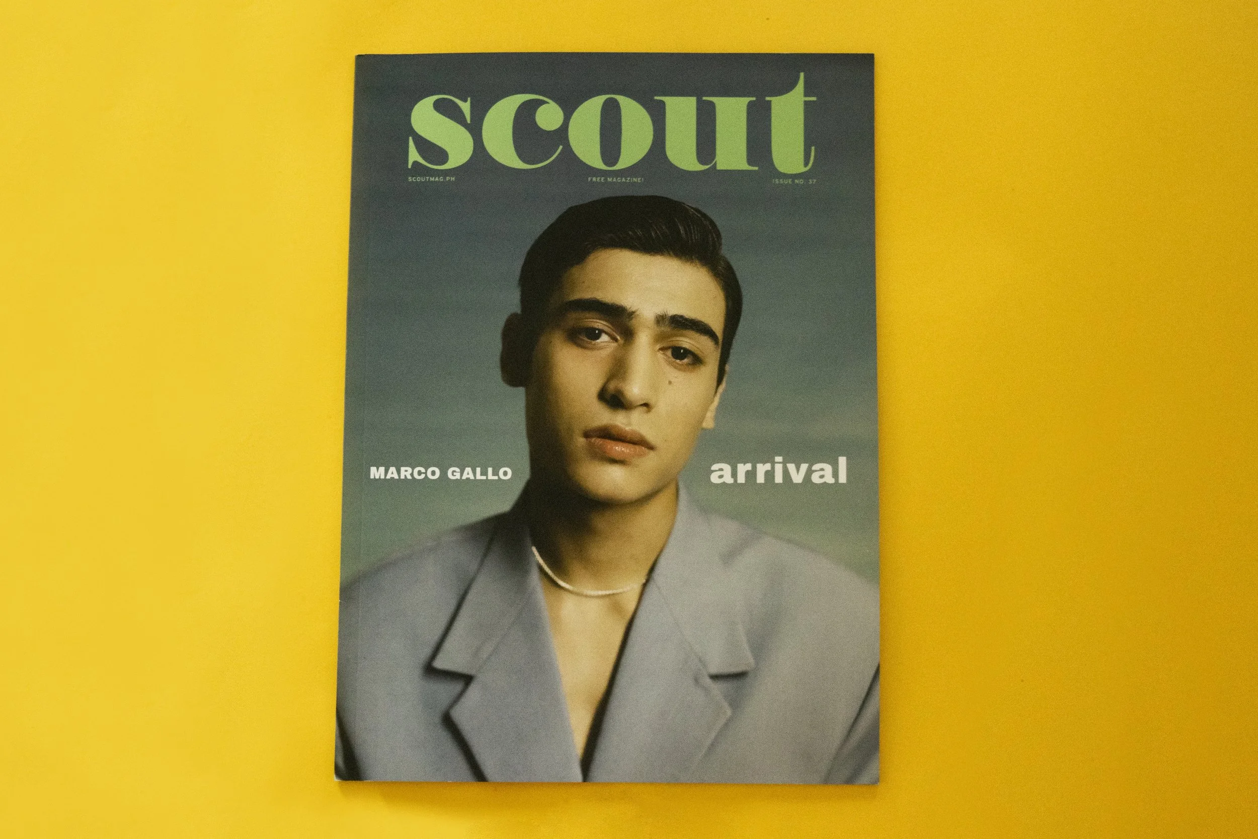 Magazine titled 'Scout' featuring a young man named Marco Gallo. He has dark hair, is wearing a blue-gray blazer. The magazine is on a yellow background.