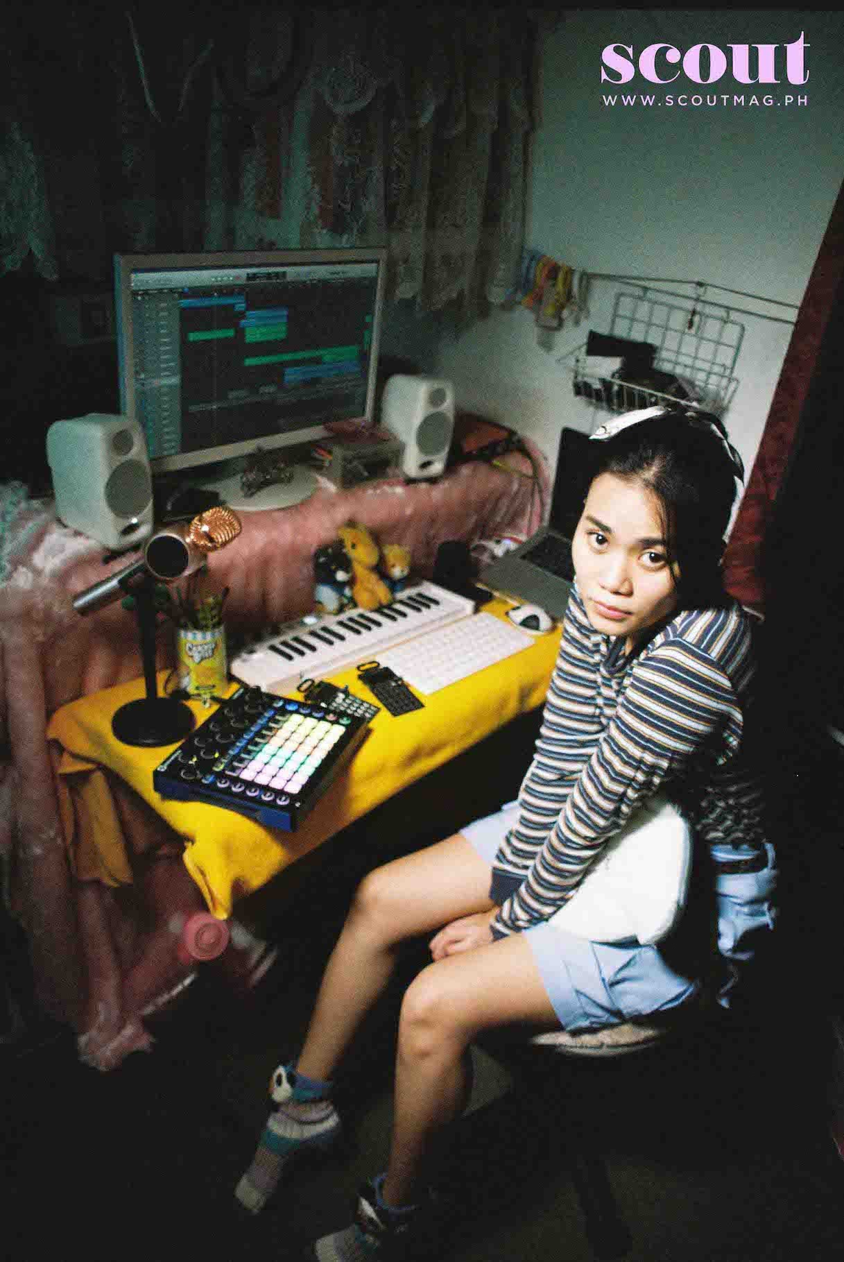 A young woman sitting in a music production room surrounded by electronic music equipment, including a computer, MIDI keyboard, and audio monitors, with a microphone and decorative items on the table beside her.