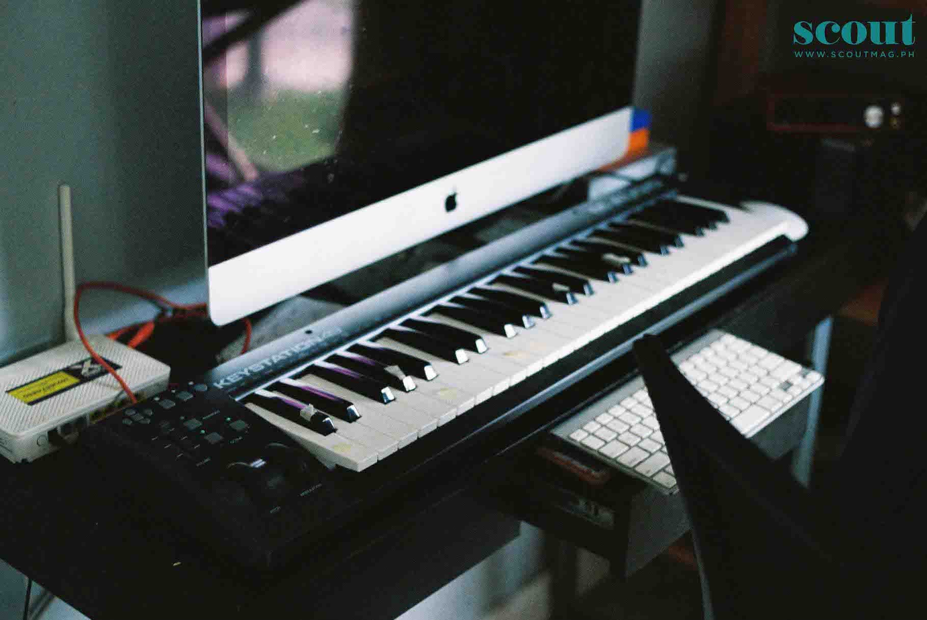 A music studio setup featuring an iMac computer placed on a desk, with a MIDI keyboard controller in front of it. To the left, there is an external audio interface with various cables attached. A wireless keyboard is placed on a chair underneath the desk, and a wireless mouse is on the desk. The image includes a logo in the upper right corner for 'Scout' with their website URL.