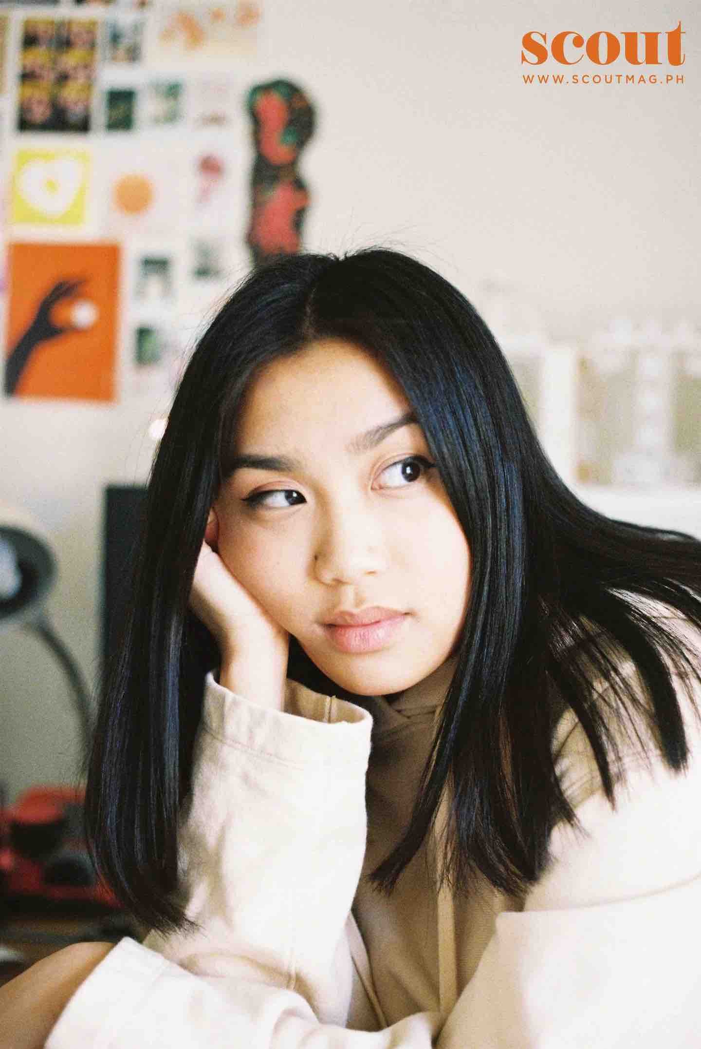 A young woman with long black hair, wearing a beige sweater, is resting her head on her hand while sitting at a desk. She is looking to her right. The background features a wall with colorful artwork and photographs, and a logo in the top right corner that says 'scout' in orange text with a website link.