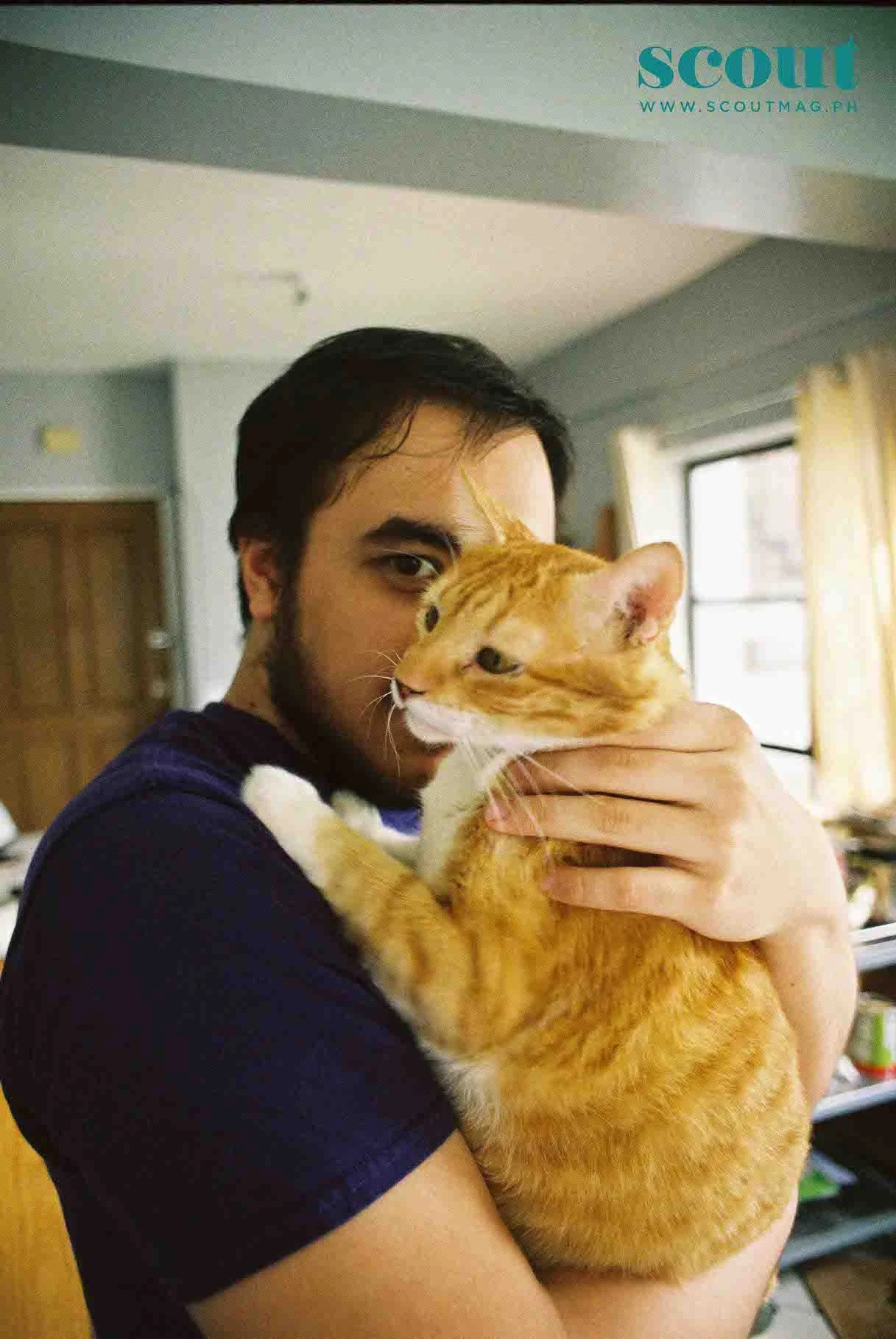 A man with black hair and a beard holding an orange tabby cat inside a home, with yellow curtains and windows in the background.