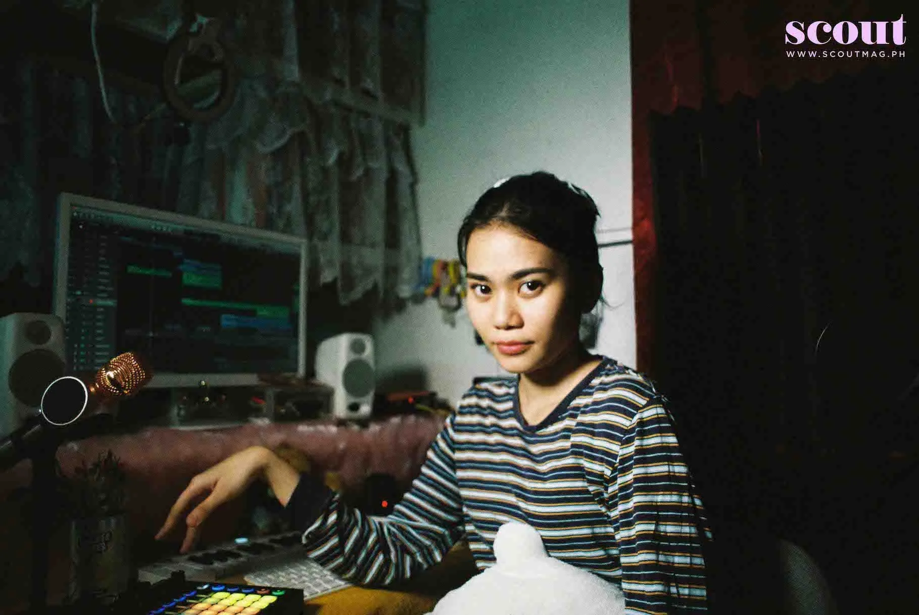 A young woman in a striped shirt sitting at a desk with music production equipment, including a computer with digital audio workstation software, speakers, a microphone, and a MIDI controller in a dimly lit room.