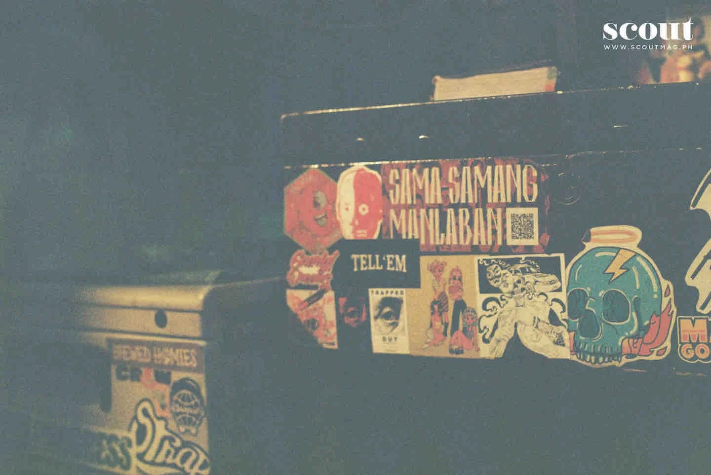Close-up of a sticker-covered black surface featuring colorful and vintage-style stickers, including a skull with a lightning bolt, a face with an eye, and various text stickers. A white sticker on the right reads "SCOUT" with a web address underneath.