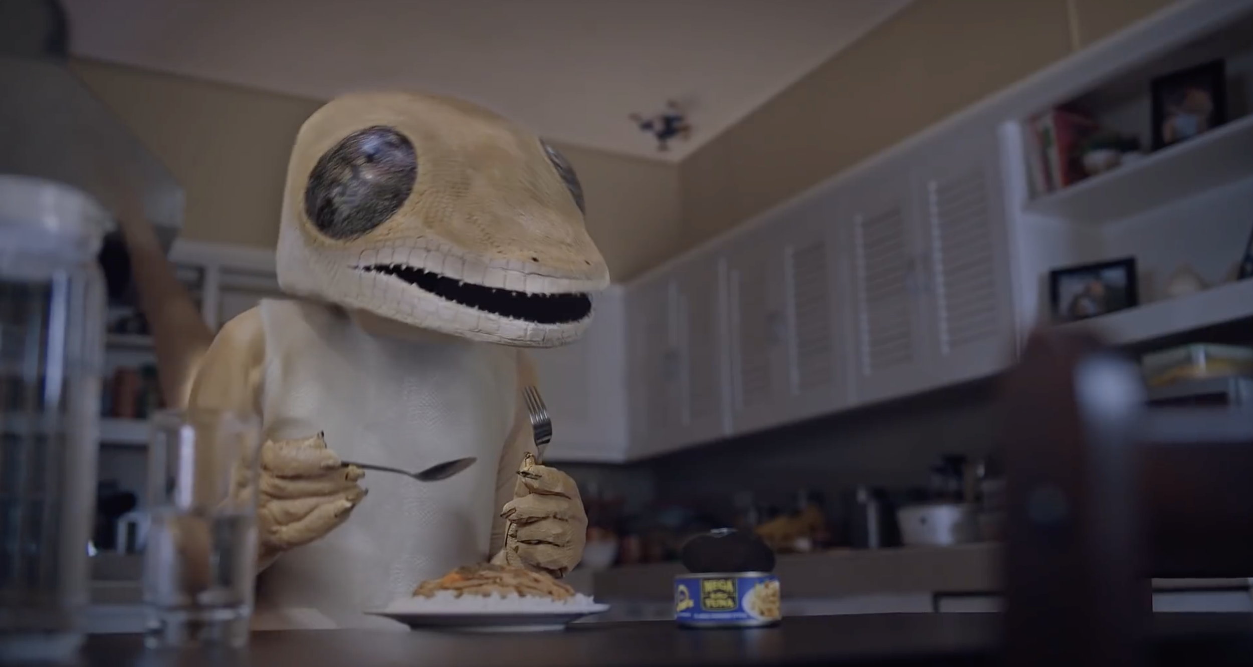 A humanoid figure with a dinosaur skull for a head, dressed in a beige outfit, sitting at a table with various food items and utensils in a kitchen.
