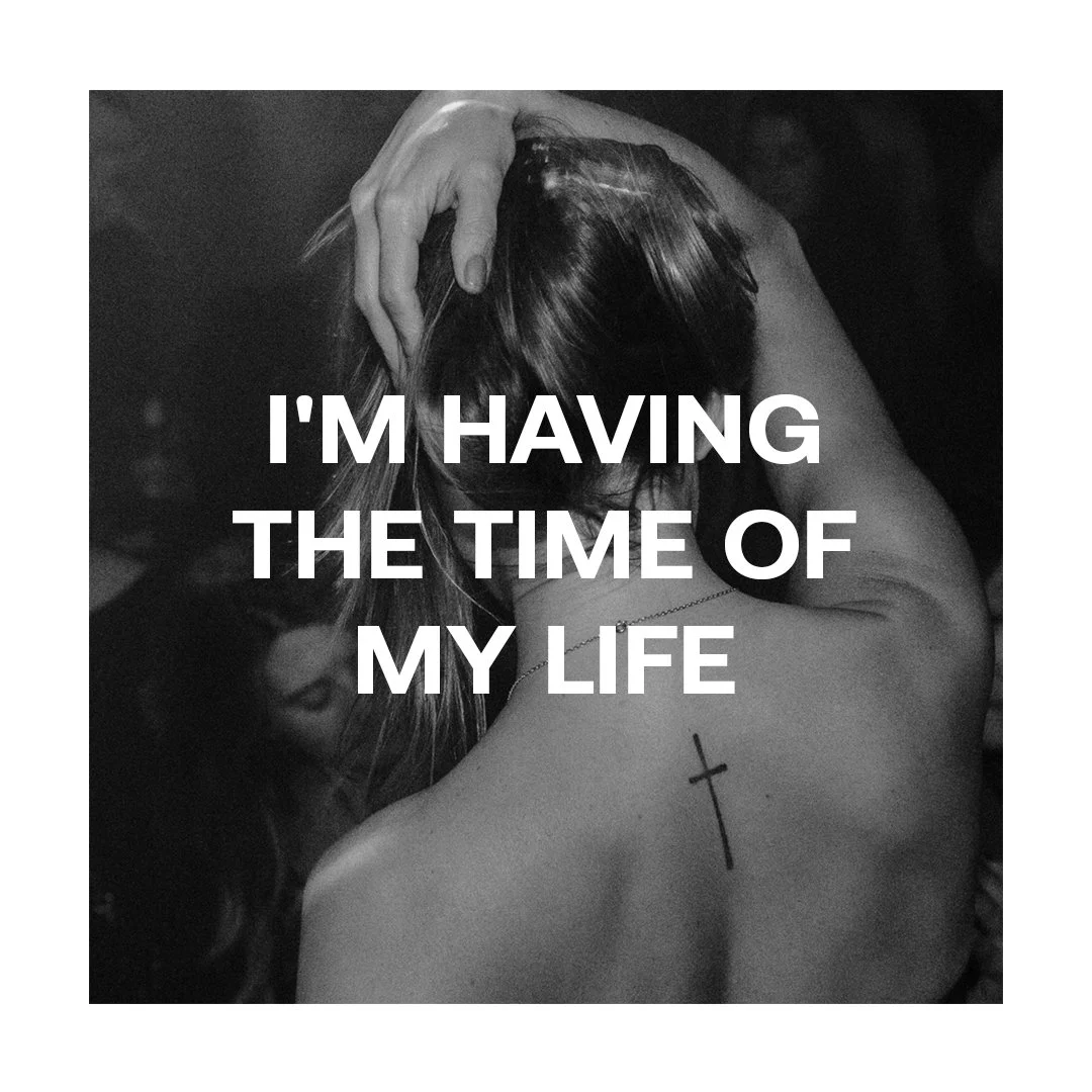 Black and white photo of a woman with her head bowed, hand on her hair, tattoo of a cross on her shoulder, overlaid with the text 'I'M HAVING THE TIME OF MY LIFE'.
