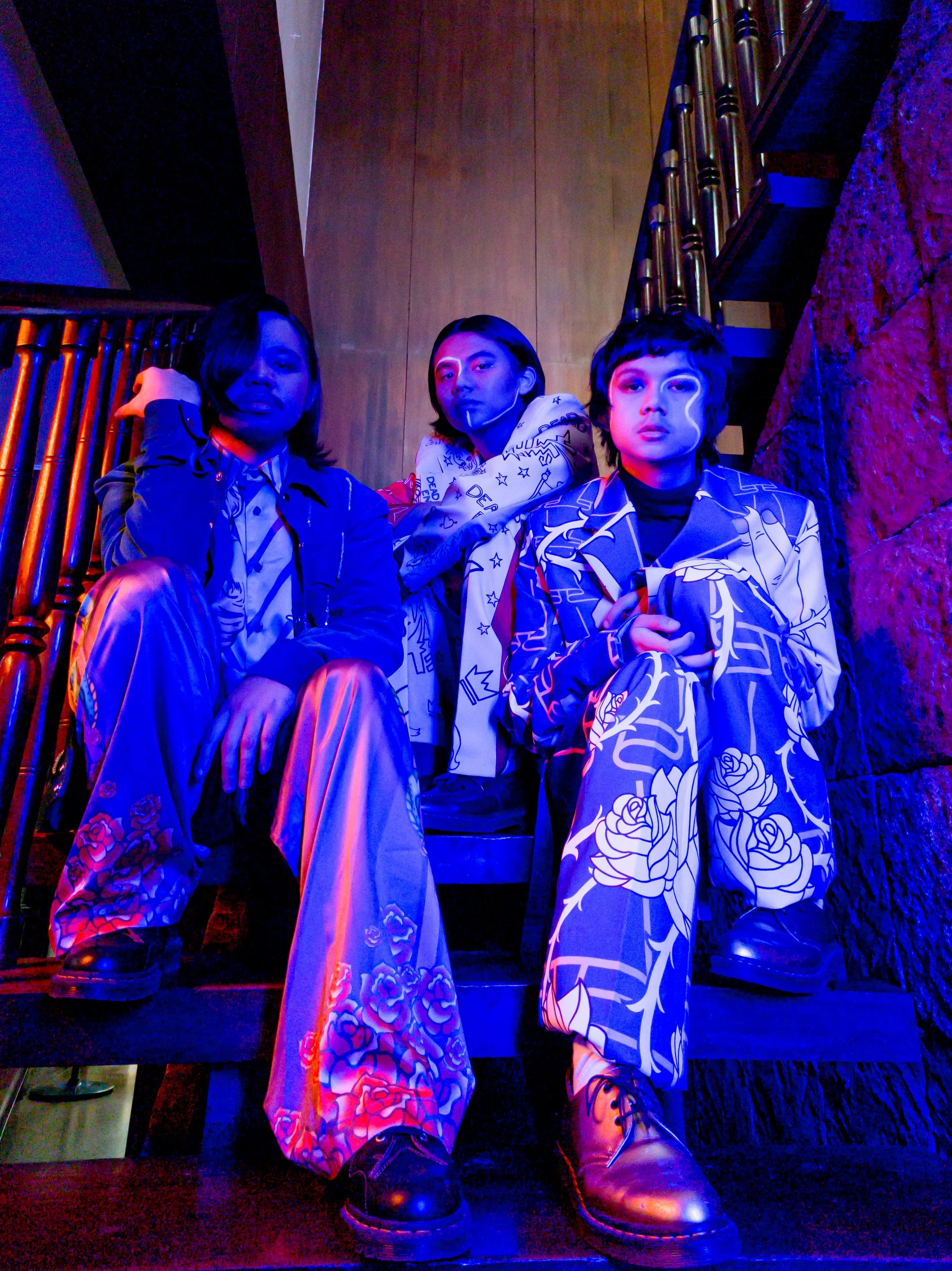 Three individuals sitting in a dimly lit room with colorful lighting, wearing patterned clothing, in front of a wooden wall and staircase railing.