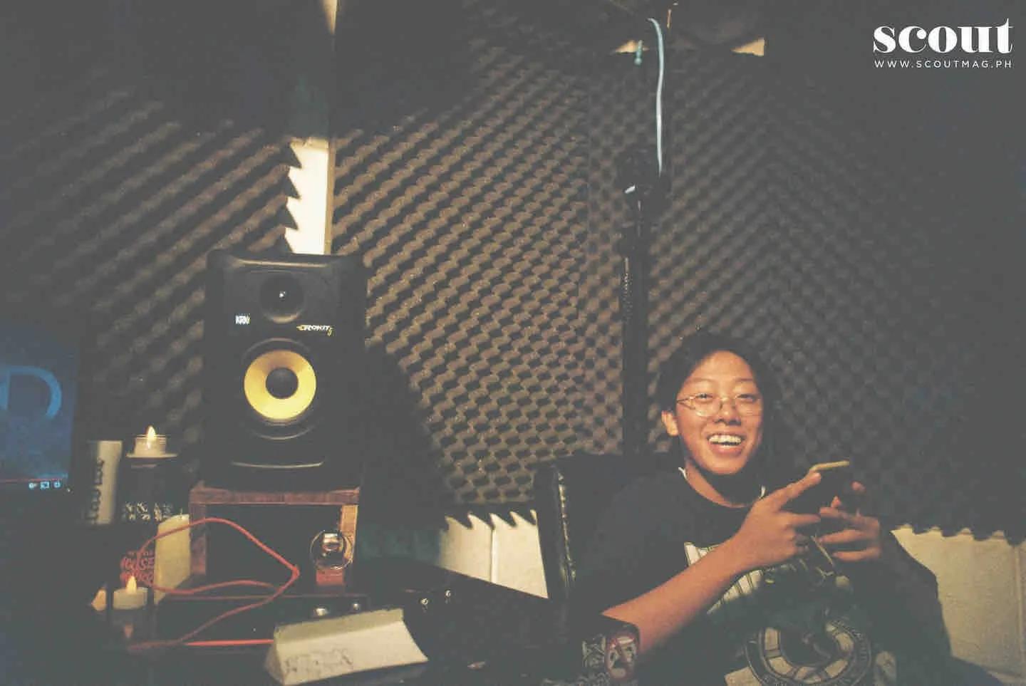 Person smiling and holding a mobile phone in a music recording studio with soundproof foam walls, studio monitor speaker, and audio equipment.