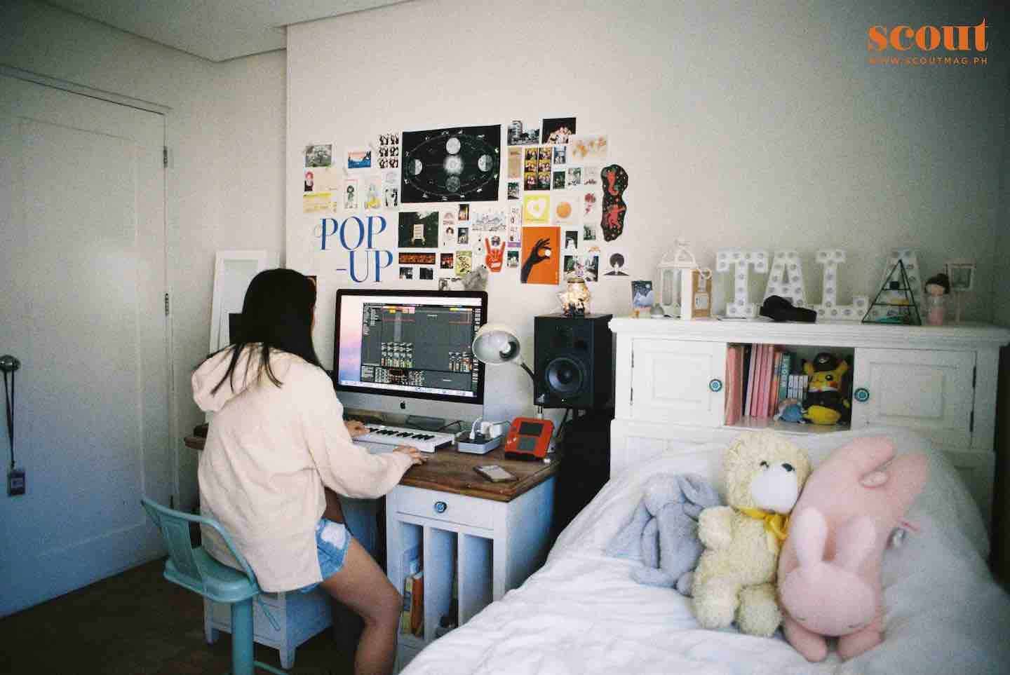 A woman sitting at a desk with a computer and speakers, decorated with posters and artwork on the wall, and plush toys on the bed next to a white cabinet in a bedroom or studio space.