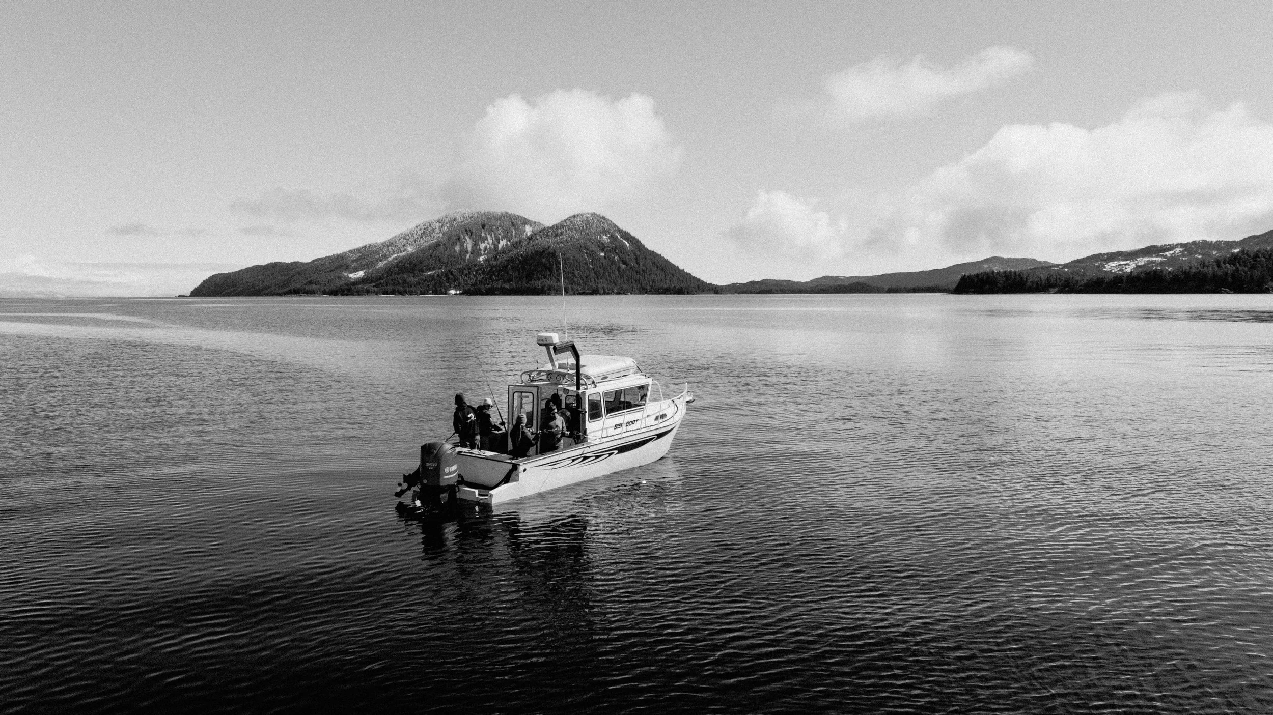 A boat with four people on board sailing in a calm body of water with mountains in the background and partly cloudy sky.