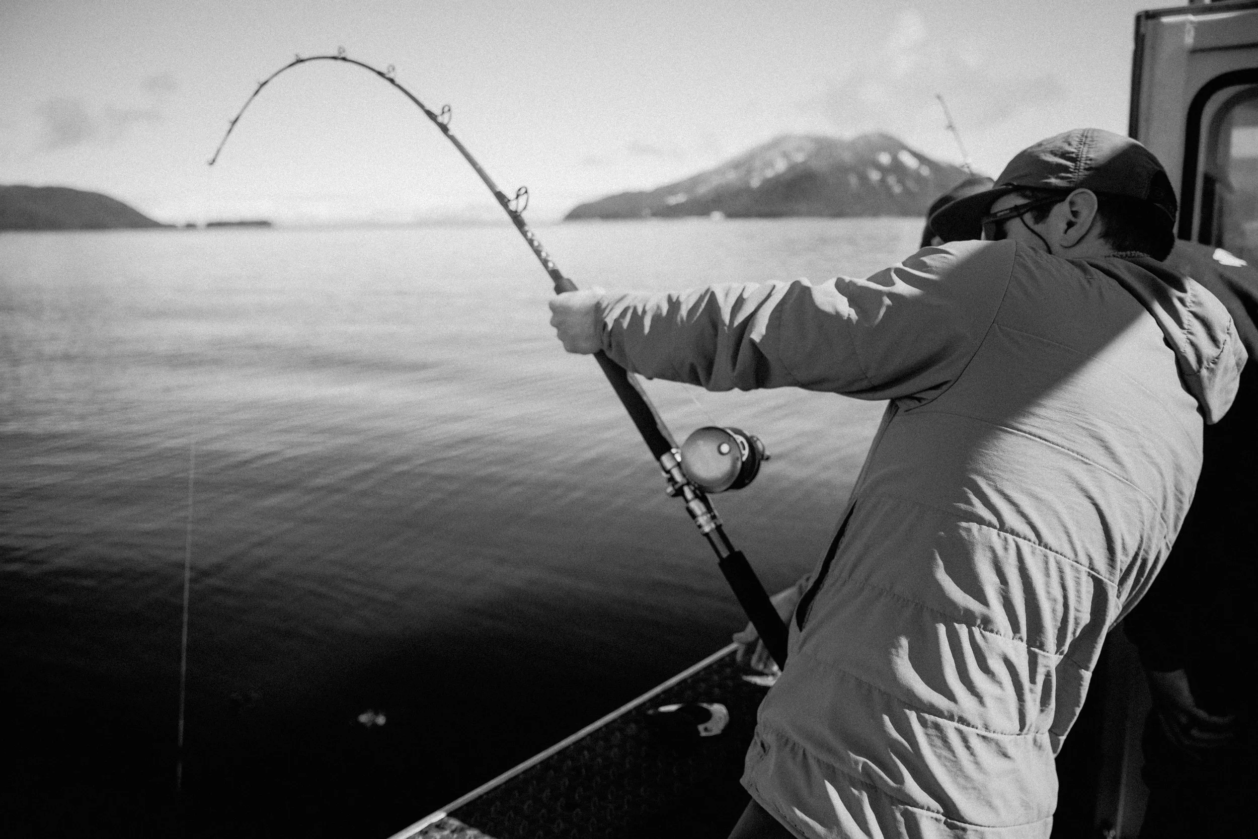 A person in a jacket and cap fishing into a body of water. The scene is in black and white with mountains in the background.