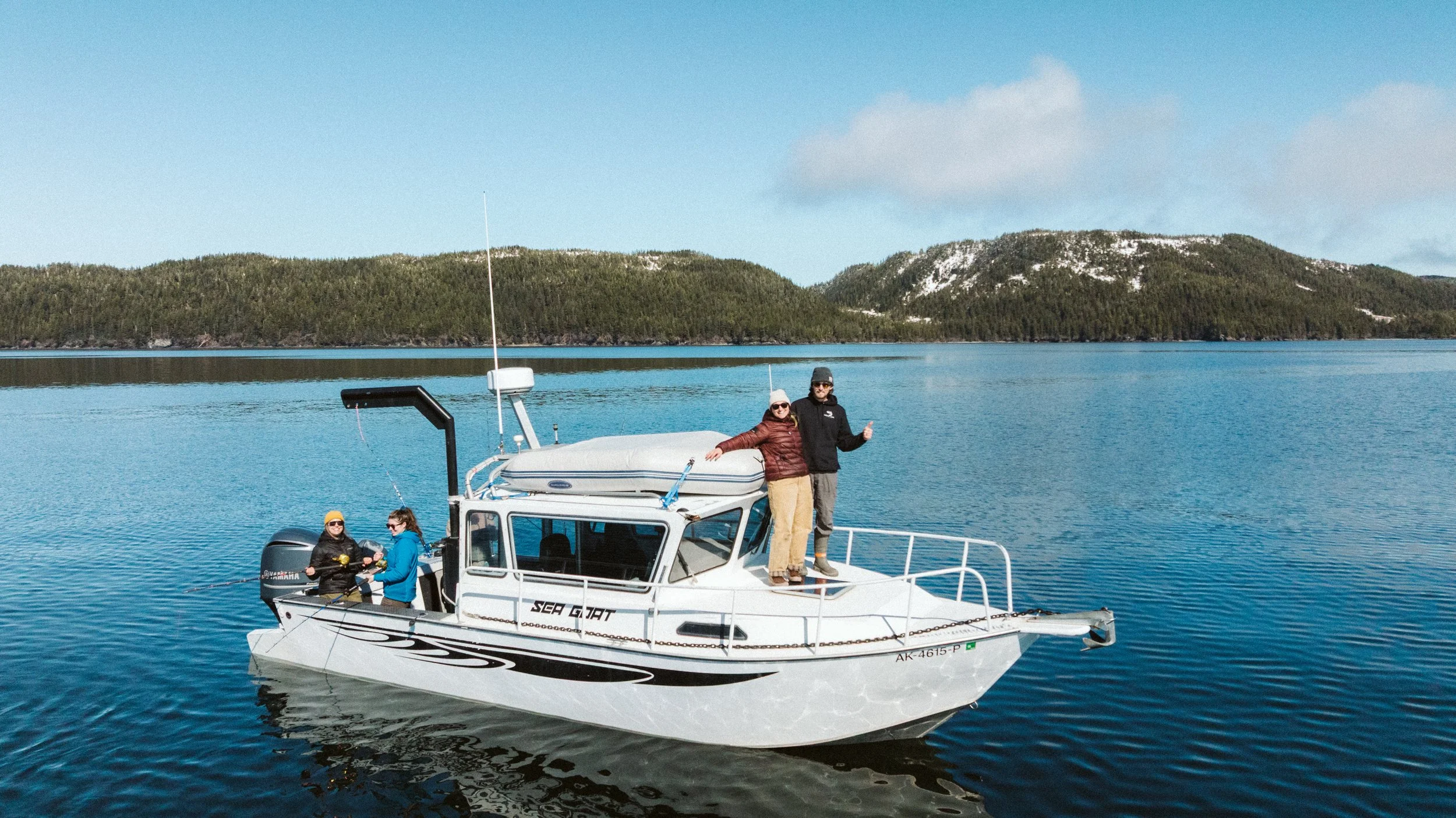 A group of people on a white boat named Sea Scout, towing a person on a fishing line in a body of water, with a forested mountain range in the background on a sunny day.