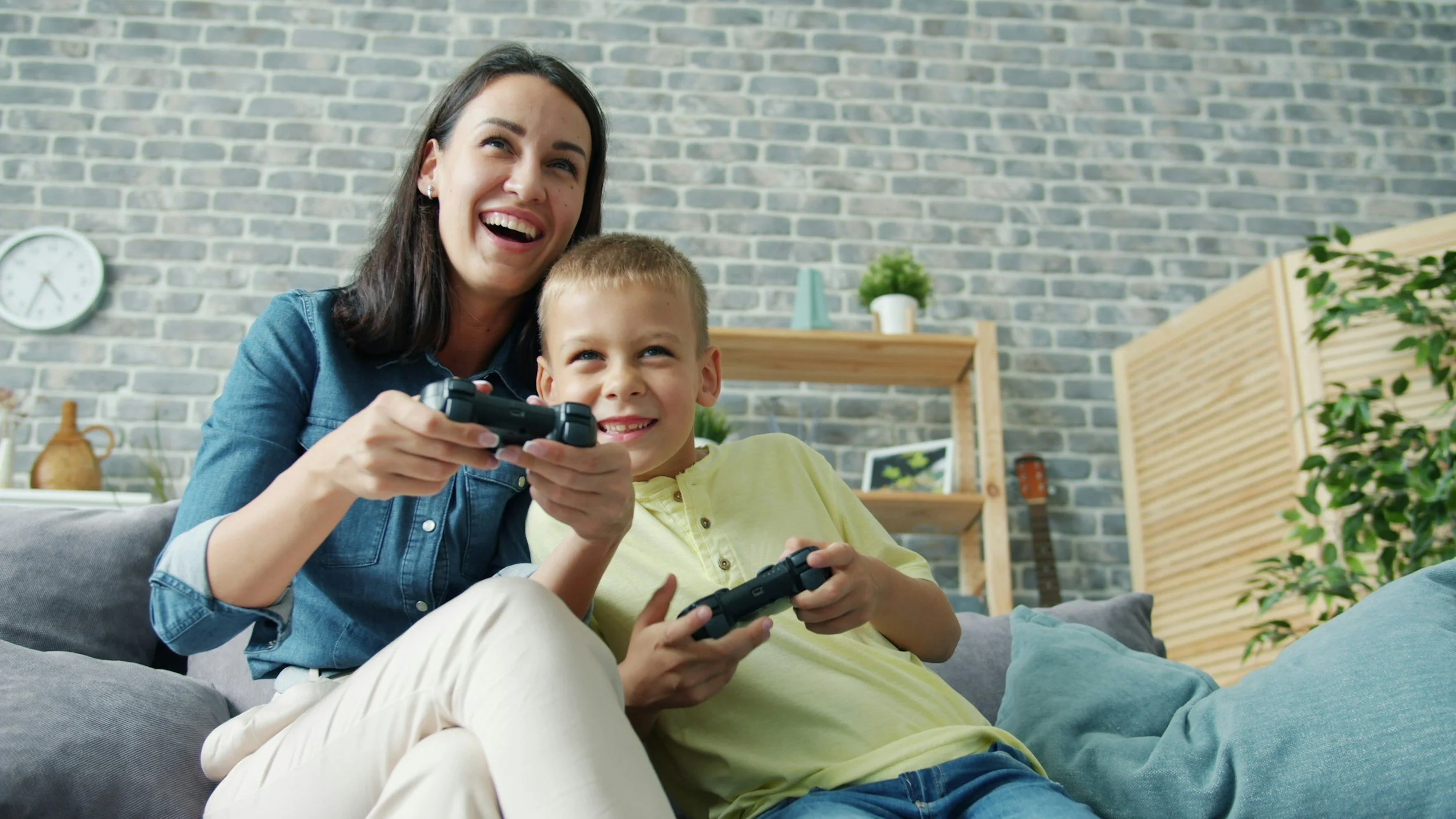 A woman and a young boy sitting on a gray sofa, playing video games together, smiling and enjoying their time.