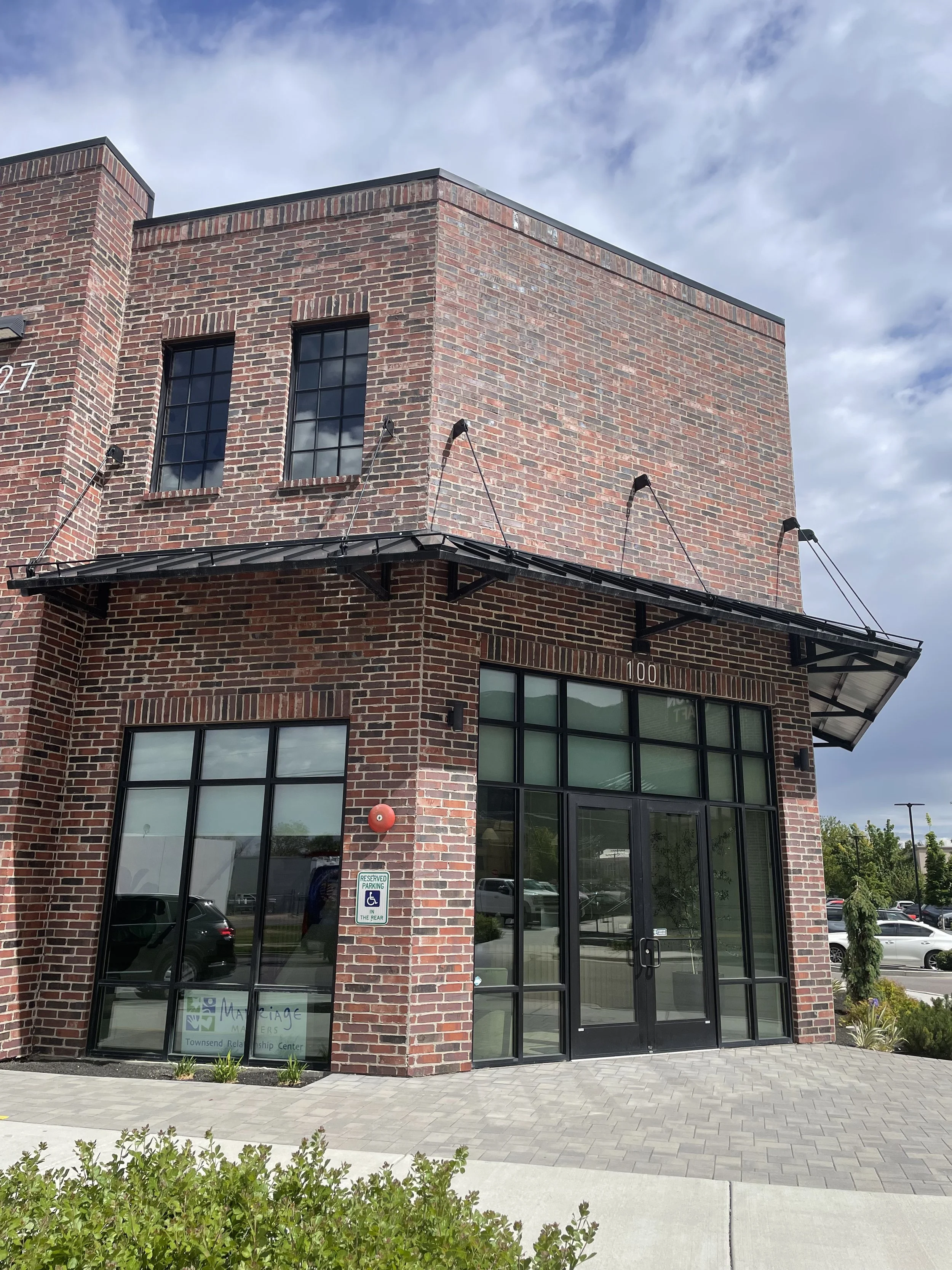 A modern brick building with large glass windows and a glass door, under a partly cloudy sky. There is a sign for a therapy center on the window, and a reserved parking sign for handicapped in front.